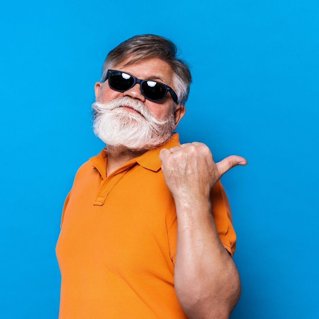 An older man with a white beard and mustache wearing black sunglasses, an orange polo shirt, and pointing to his shoulder against a blue background.