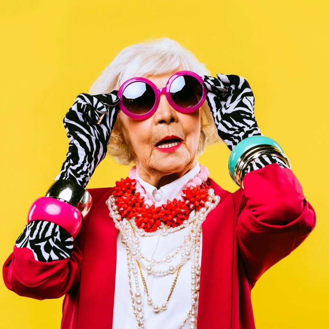 Older woman with white hair wearing large pink sunglasses, black and white zebra print gloves, a red blazer, and multiple strands of beads and necklaces, posing against a yellow background.