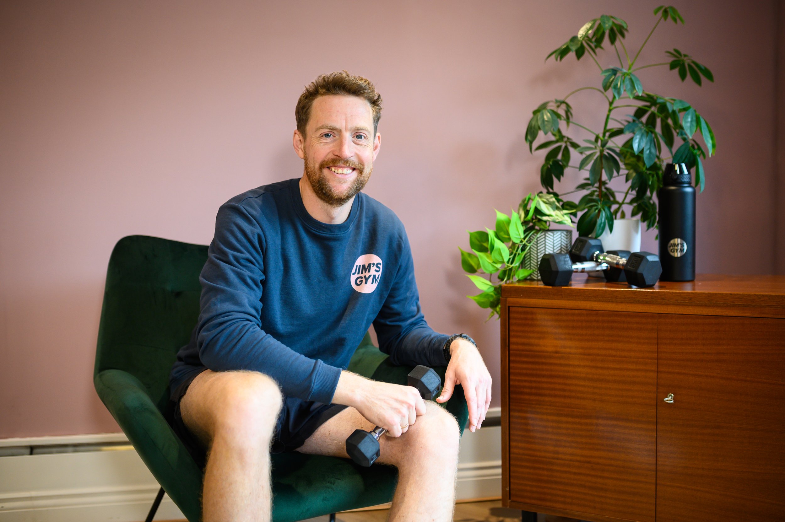 A man sitting in a green chair smiling, wearing a blue sweatshirt that says 'Jim's Gym', lifting dumbbells in a room with a pink wall, a wooden cabinet, and potted plants.
