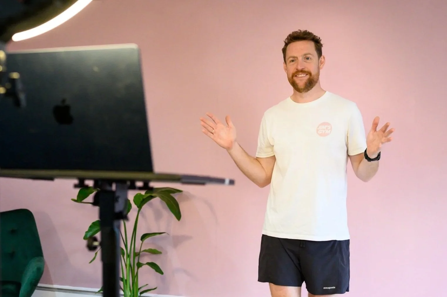 A smiling man with curly hair and a beard sitting on a workout bench, wearing a black t-shirt, black shorts, white sneakers, a black watch, and posing in a gym.