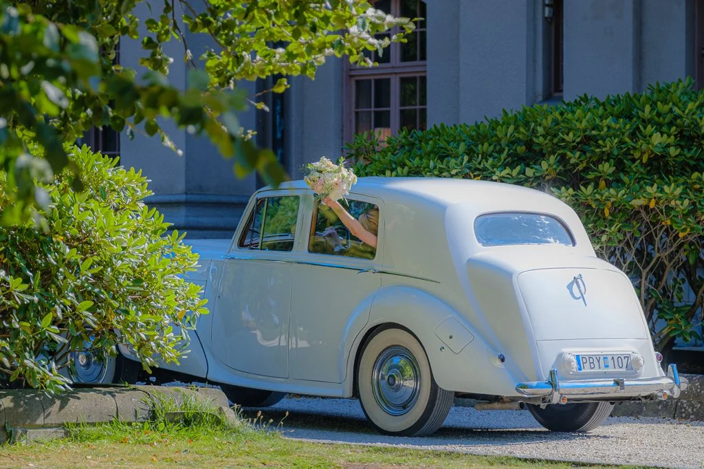 En vit vintagebil parkerad vid ett hus, med en person som sträcker sig ut ur passagerarsidan och håller i en bukett blommor.