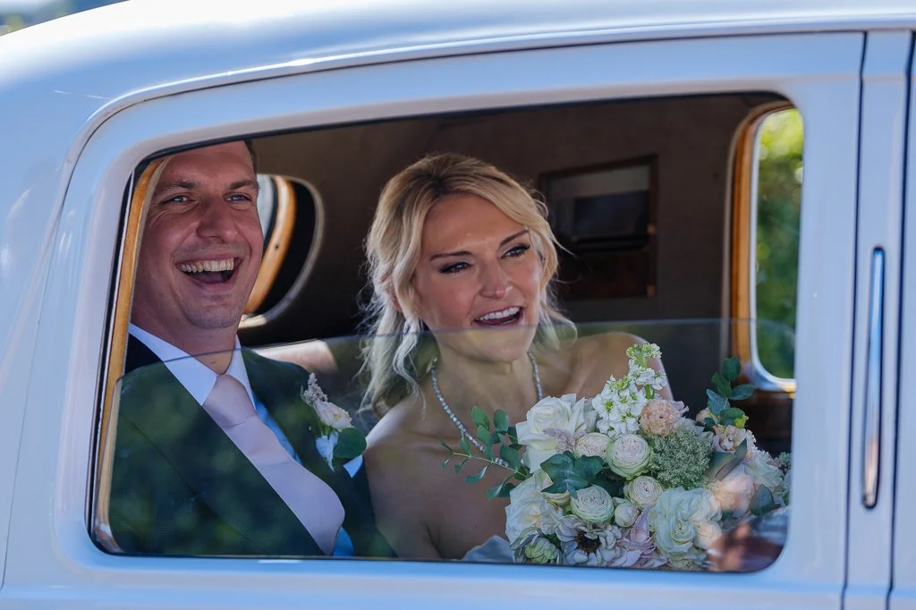 En brud och brudgum, sitter i en vit vintagebil. Kvinnan håller en stor bröllopsbukett. Bilen har en bakfönster som ger en klar vy av dem.