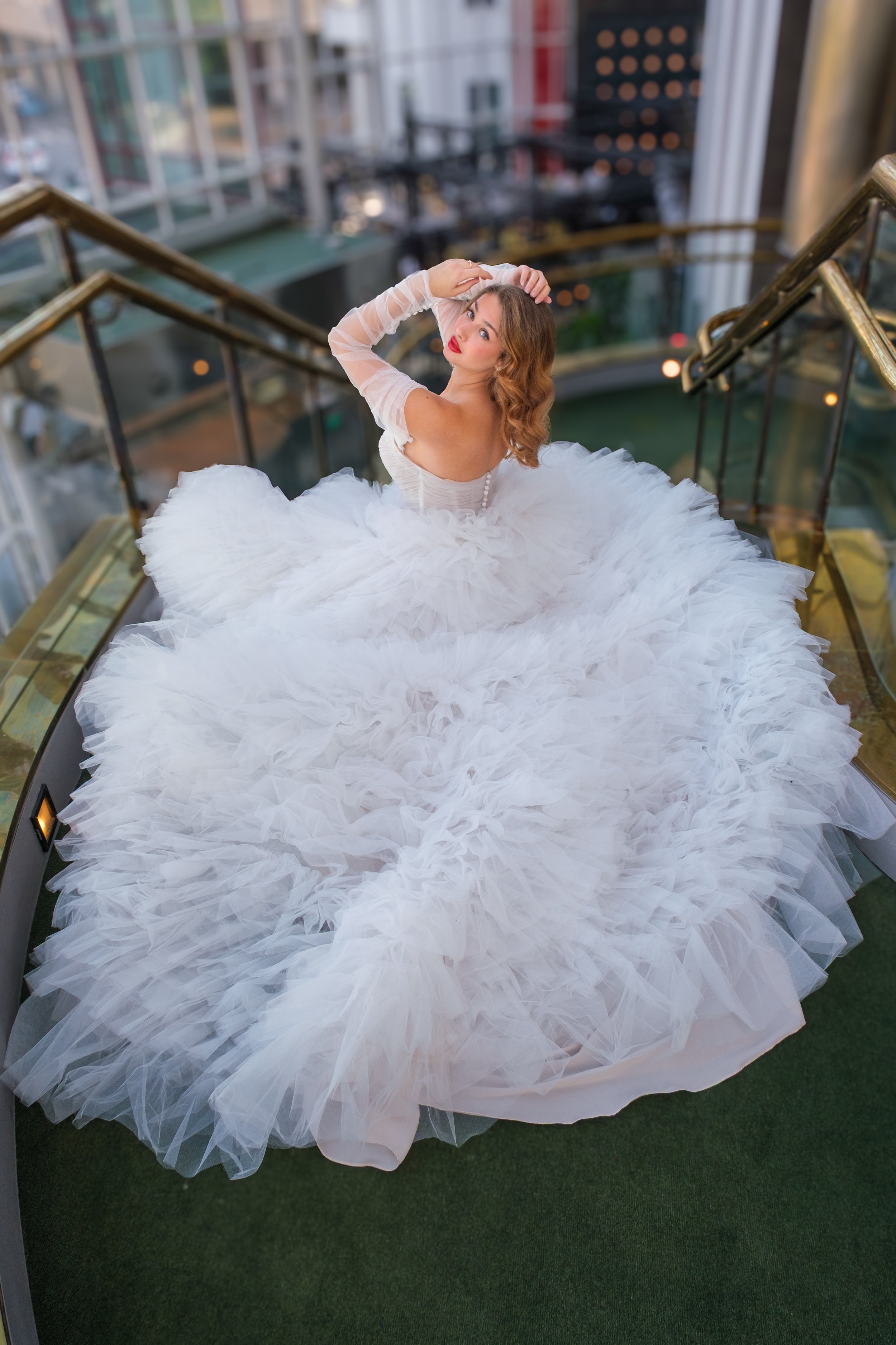 En kvinna i en vit brudklänning sitter på en spiraltrappa på en balkong med stadsvy i bakgrunden.