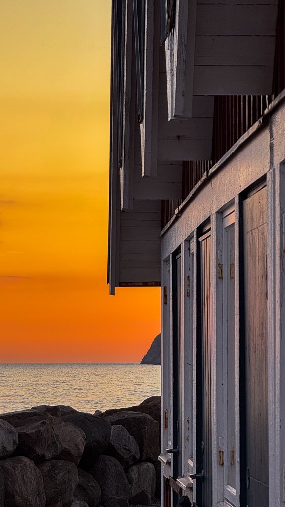 En fyrkantig träbyggnad vid kusten med fönster och dörrar, Och en stenmur framför, vid solnedgång eller soluppgång, med havet i bakgrunden och en brant klippa i horisonten.