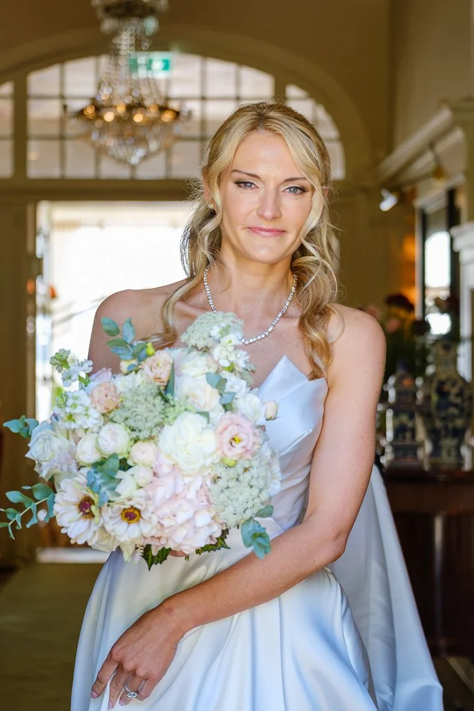 En brud i vit brudklänning håller en stor bukett med vita och ljusrosa blommor, i ett elegant inrett rum.