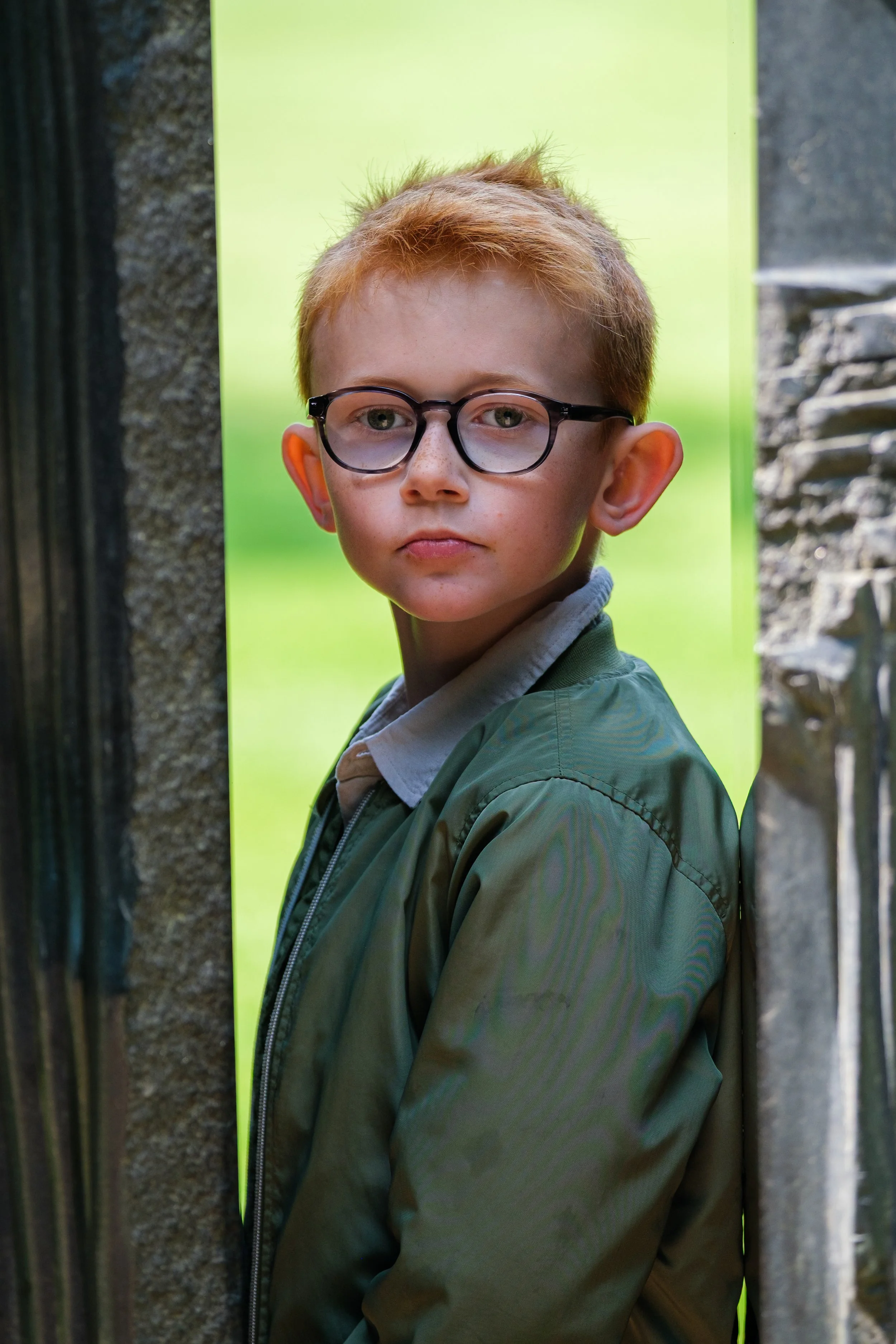 Ung pojke med glasögon står mellan en stenvägg och en svart järngrind, baksidan är en ljus grön vägg.