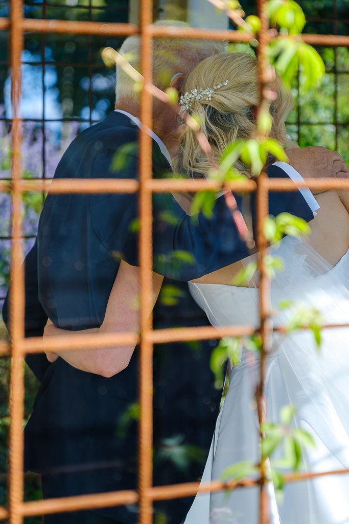 Två personer som delar ett ömt ögonblick, fotograferat genom ett metallstaket med gröna vinstockar i förgrunden.