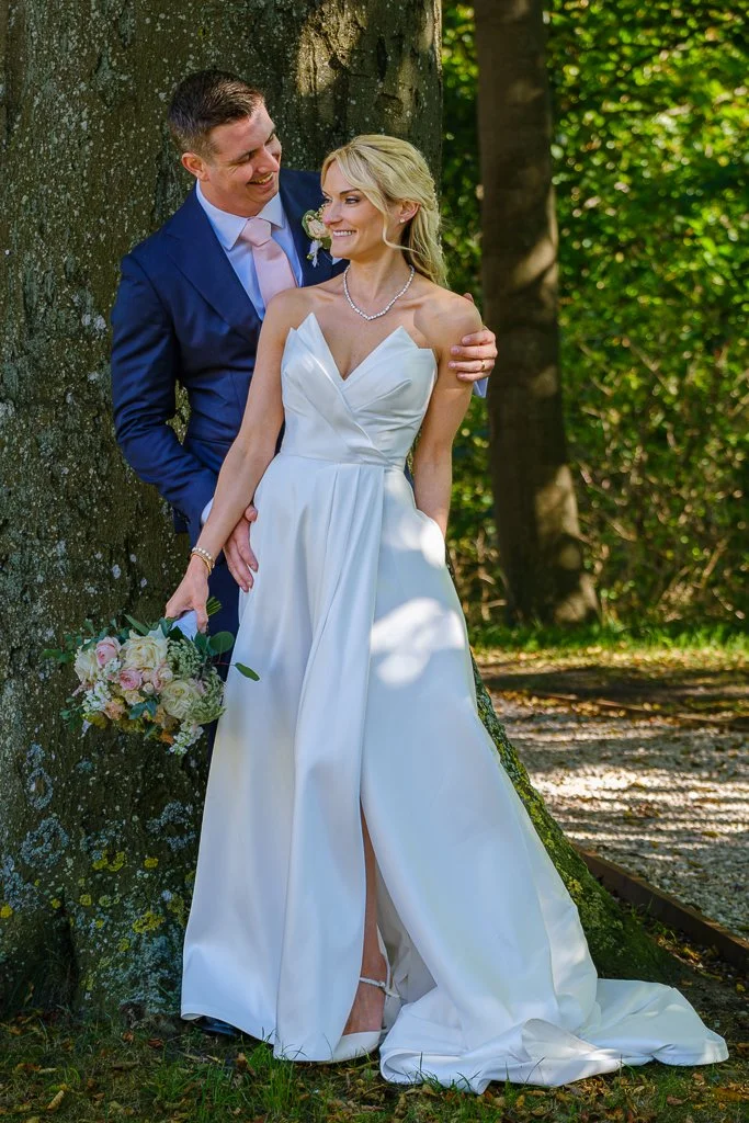 En nygift brud och brudgum står tillsammans utomhus vid ett träd, båda ler och verkar lyckliga, bruden håller en bukett blommor.