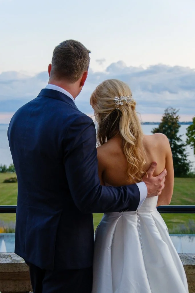 En man i mörk kostym och en kvinna i vit brudklänning står nära varandra och tittar ut över ensjölandskap i solnedgången.