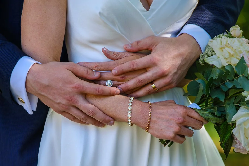 Närbild av ett äktenskapspares händer som omsluter en brud och en brudgums händer, med bröllopsring och armband, och ett bröllopsblomsterarrangemang.