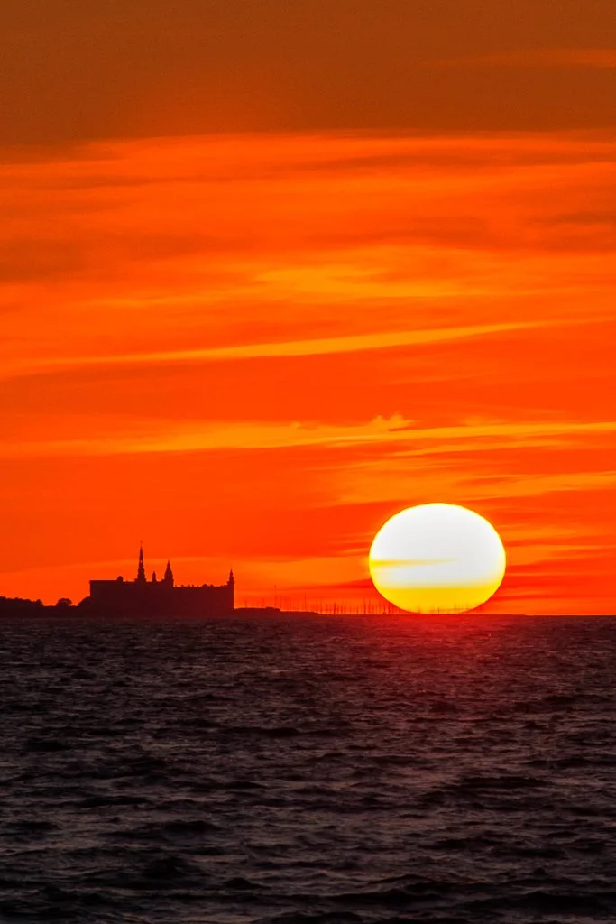 Solsnedgång över havet med en distinkt byggnad eller slott i horisonten, himlen är fylld av orange och röda nyanser.