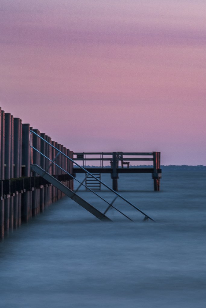 En brygga som når ut i ett lugnt hav, under en rosa och lila himmel.