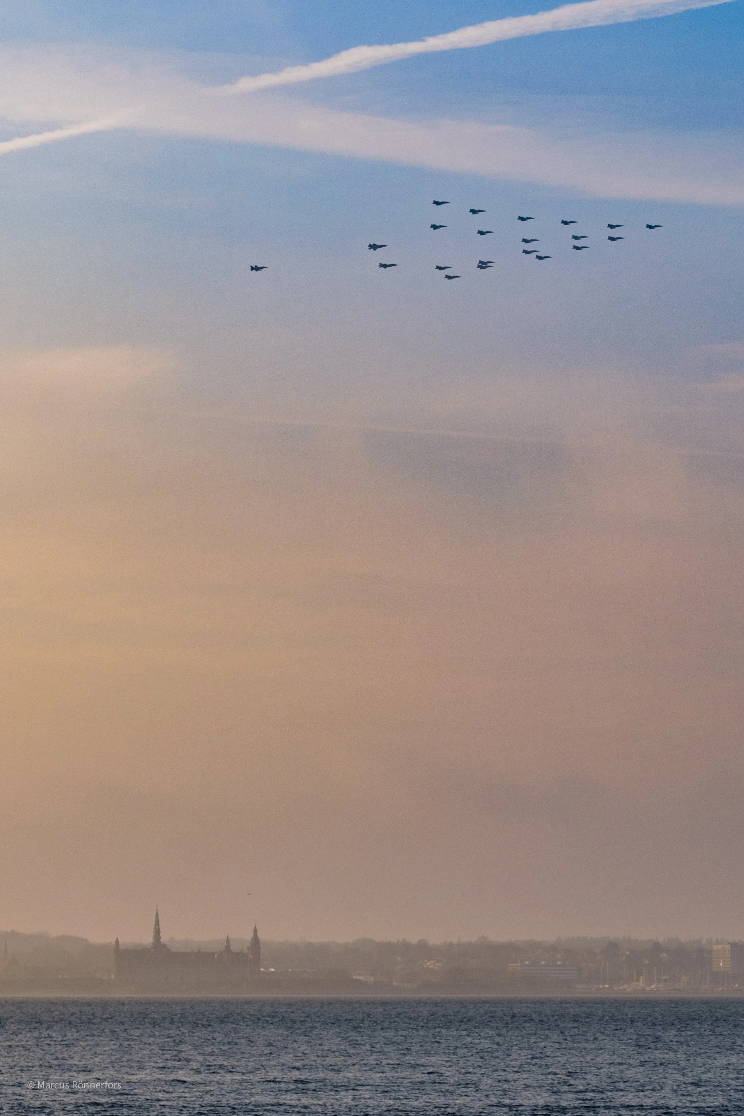 En bild av en stad vid vattnet med två kyrktorn i bakgrunden, och en flygplansformation som flyger över himlen.