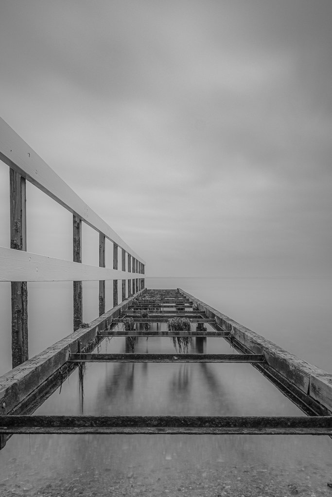 Svartvitt foto av en gammal, övergäten brygga som sträcker sig ut i vattnet under en molnig himmel.