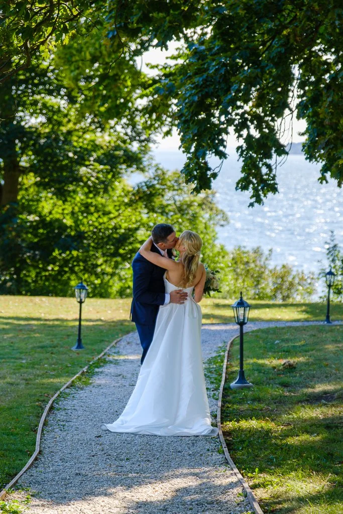 Nygifta par som kramas och kysser på en stigen vid vattnet, omgiven av gröna träd och utomhusbelysning.