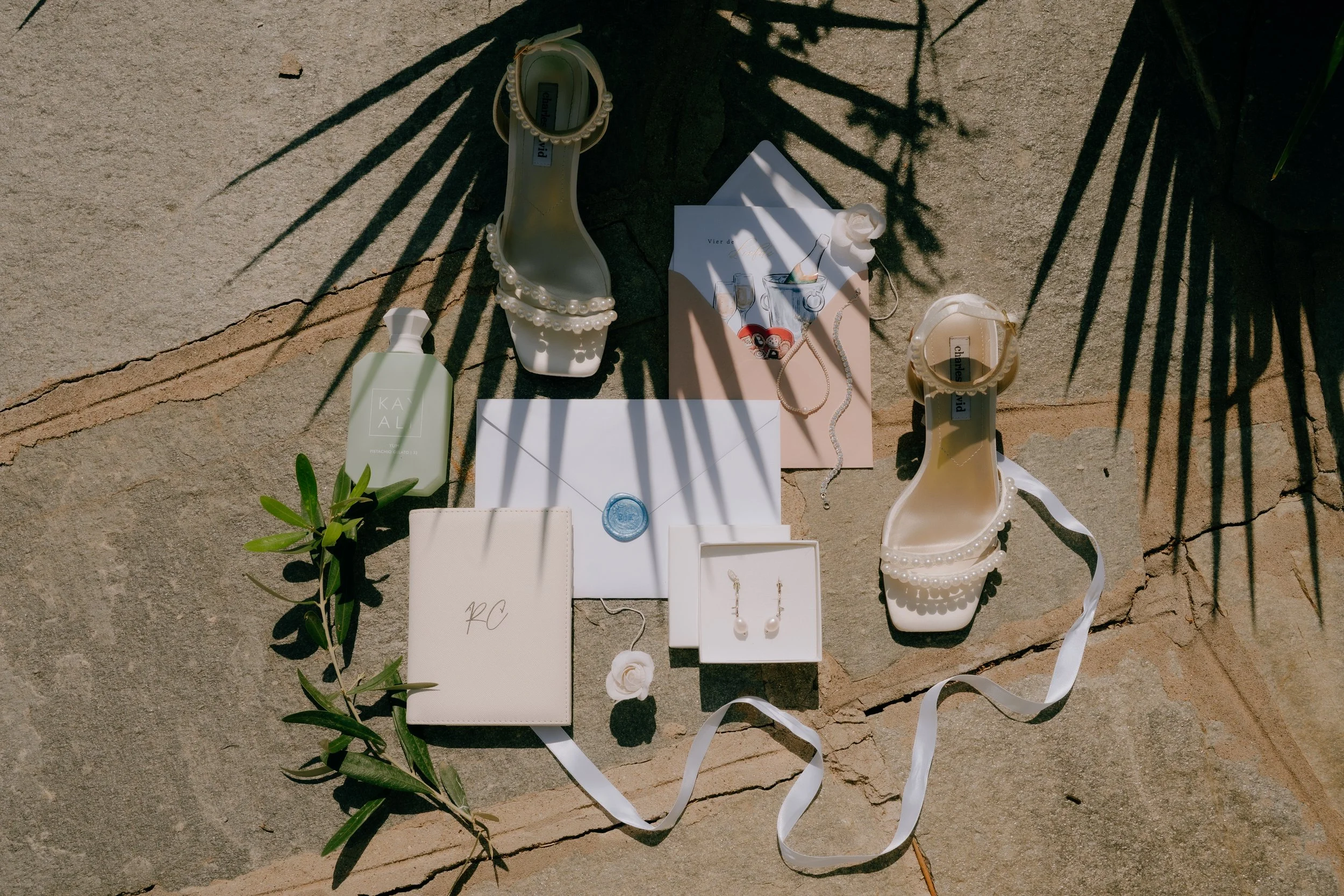 Flat lay of jewelry, shoes, and accessories on a stone surface, with shadows of palm leaves cast over them.