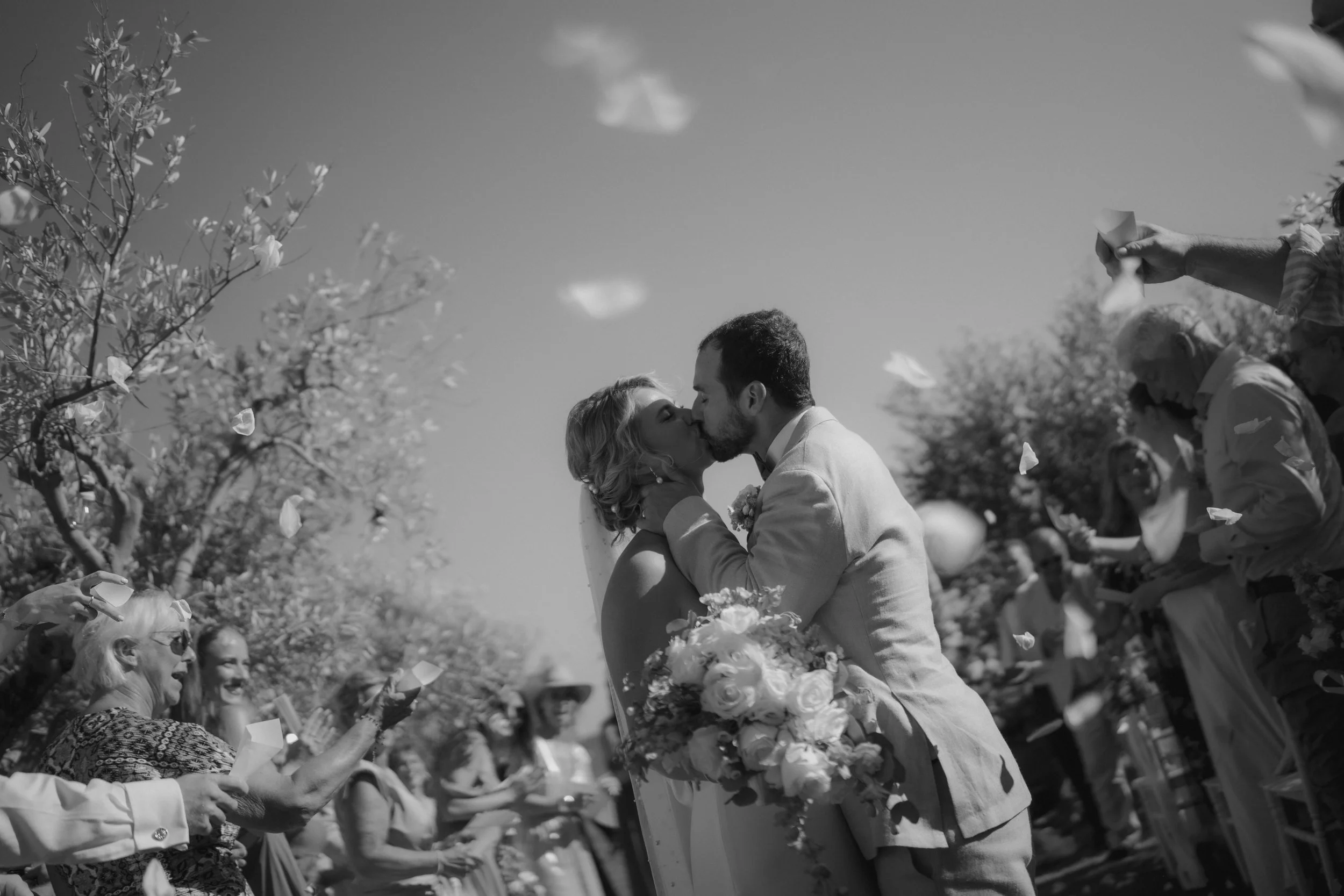 A black-and-white photo of a wedding couple kissing outdoors, surrounded by guests throwing petals and celebrating. The bride holds a bouquet of flowers, and the groom is embracing her.