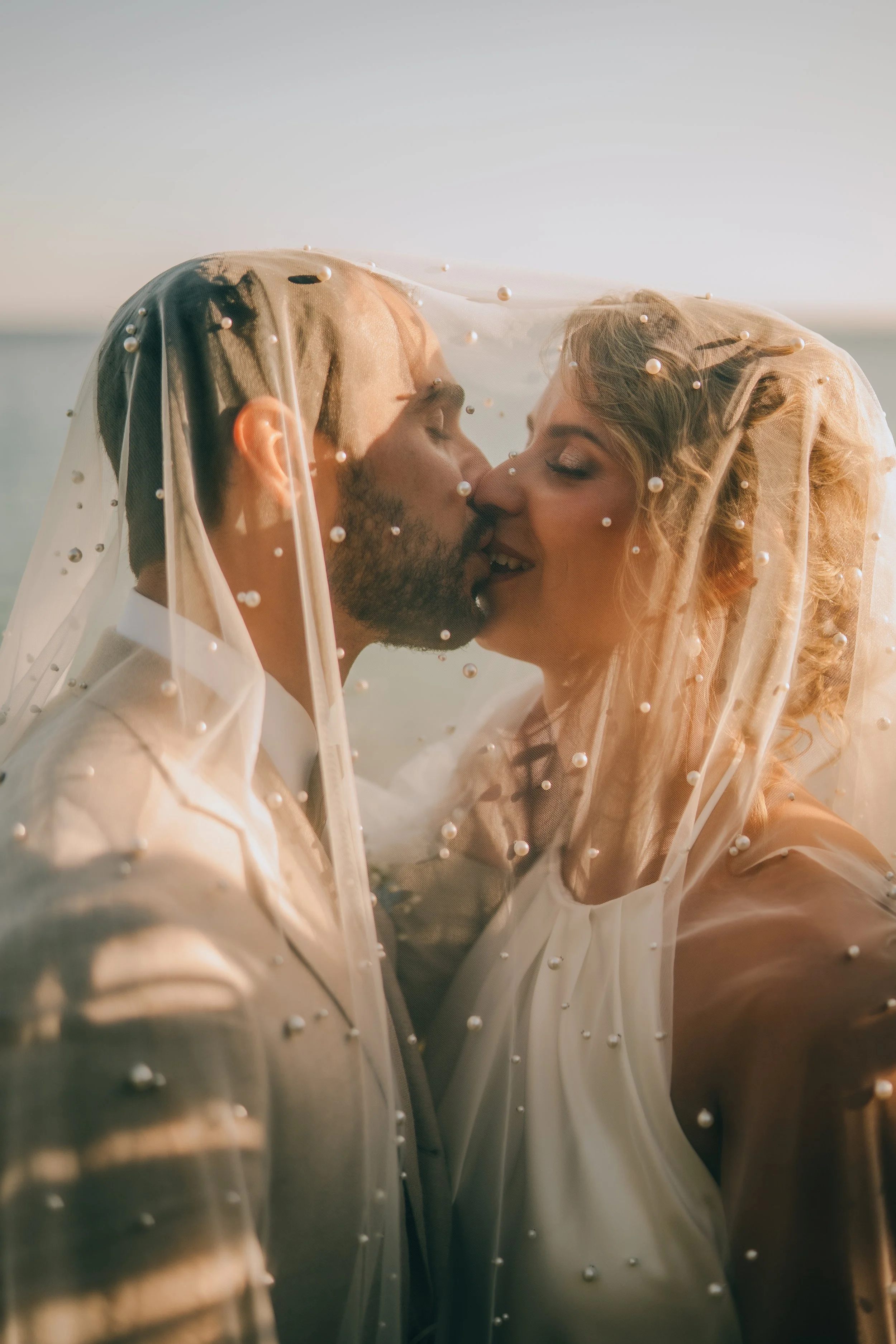 A bride and groom sharing a kiss under a veil decorated with pearls during their wedding on the beach at sunset.