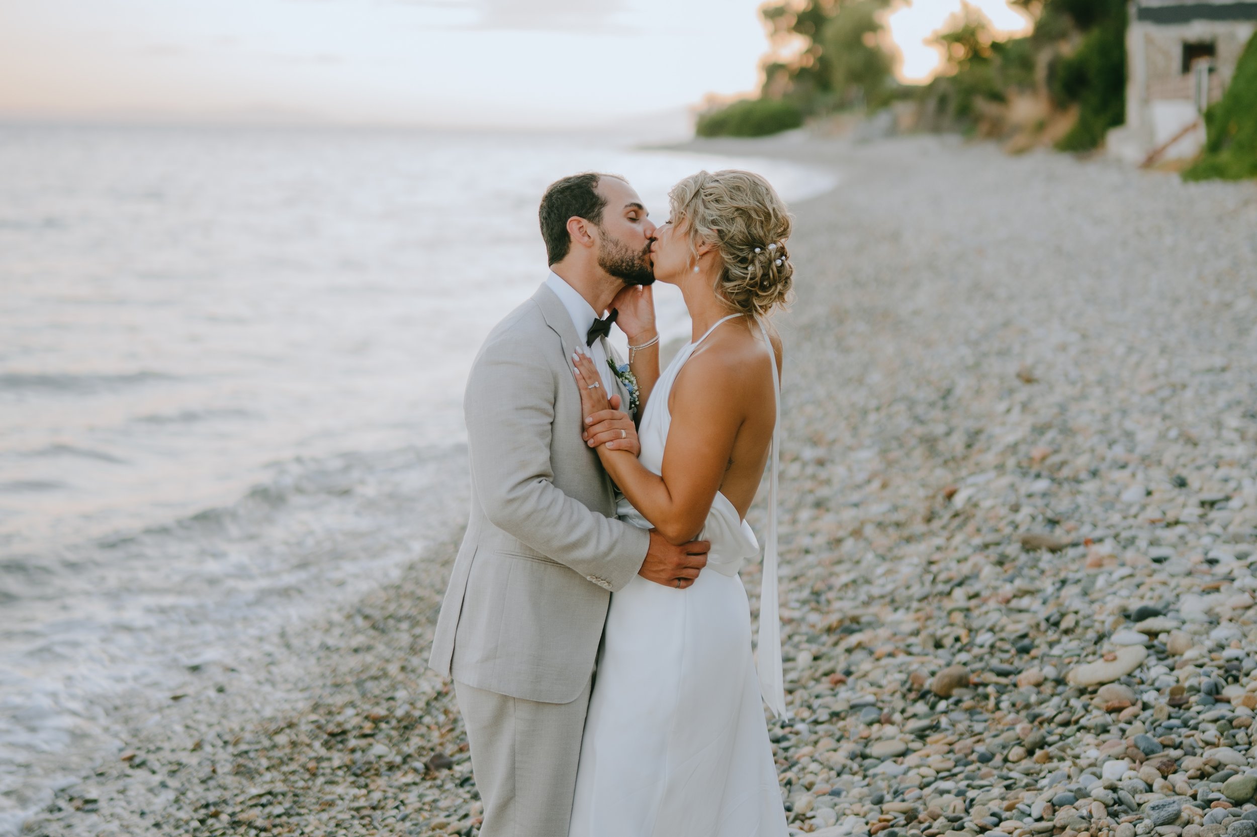 A couple in wedding attire sharing a kiss on a pebble beach at sunset.