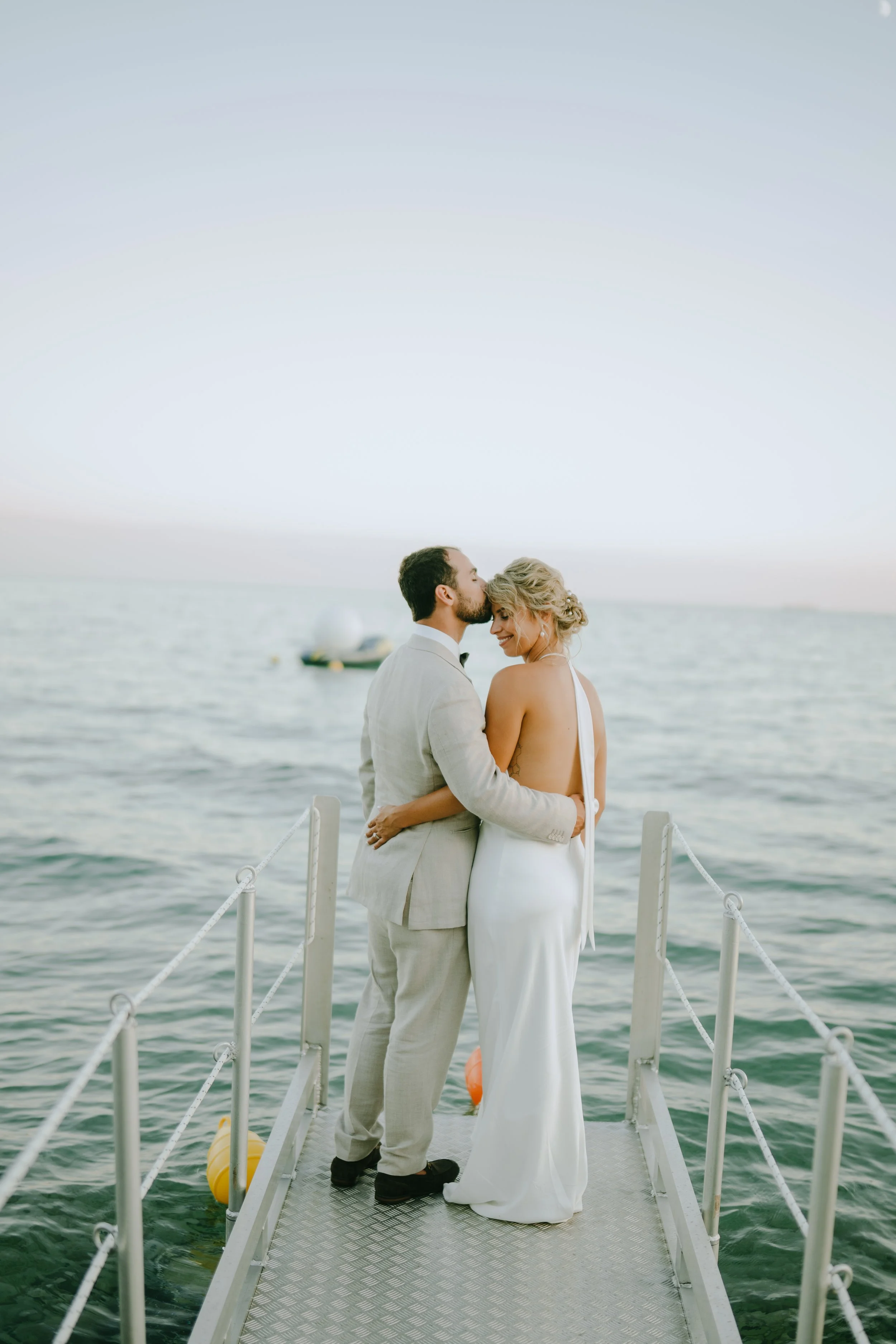 A newlywed couple embracing on a dock by the lake, with a boat in the background during sunset.