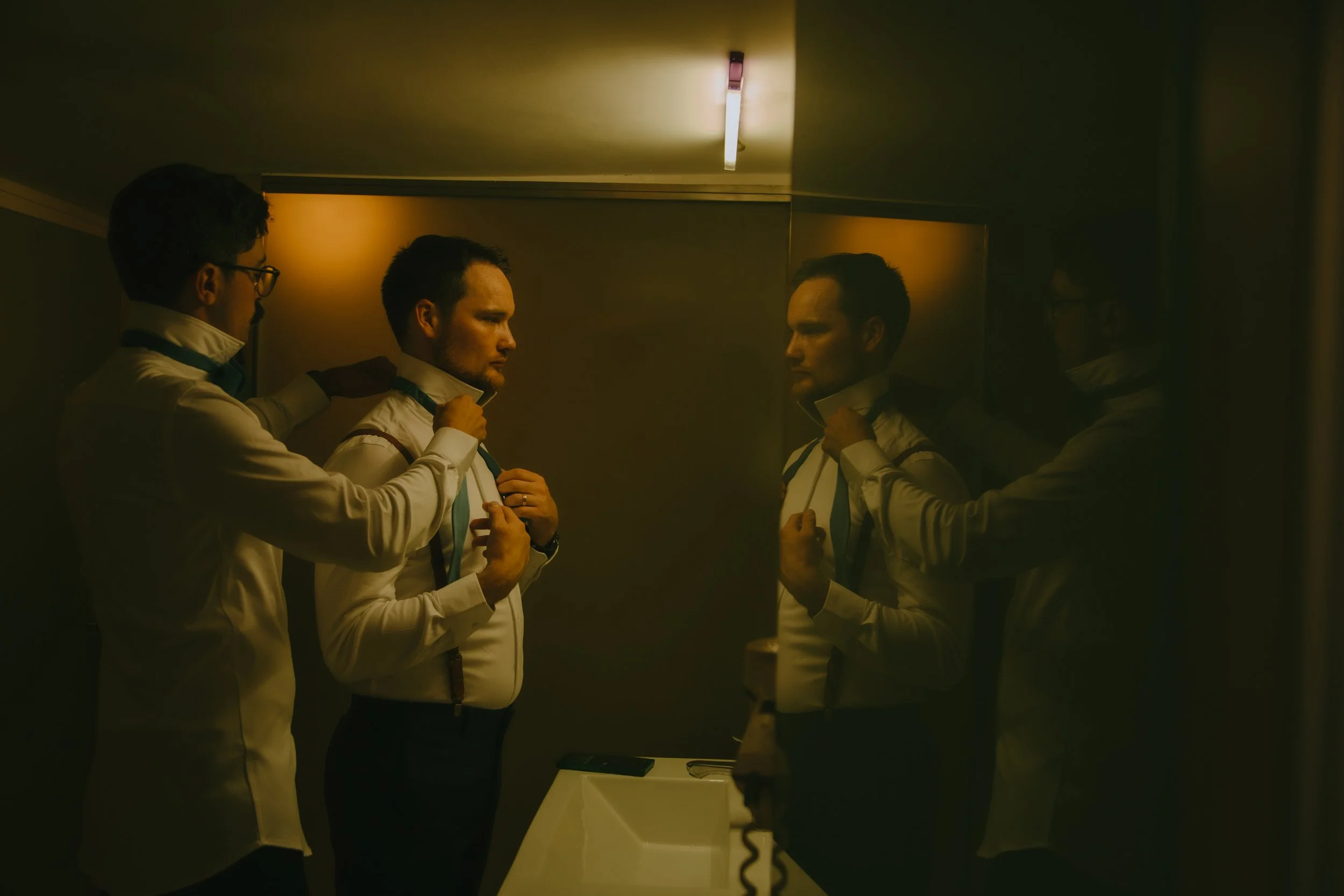 A man standing in front of a mirror adjusts his collar while two others help him, all dressed in white shirts, in a dimly lit room with yellowish lighting.