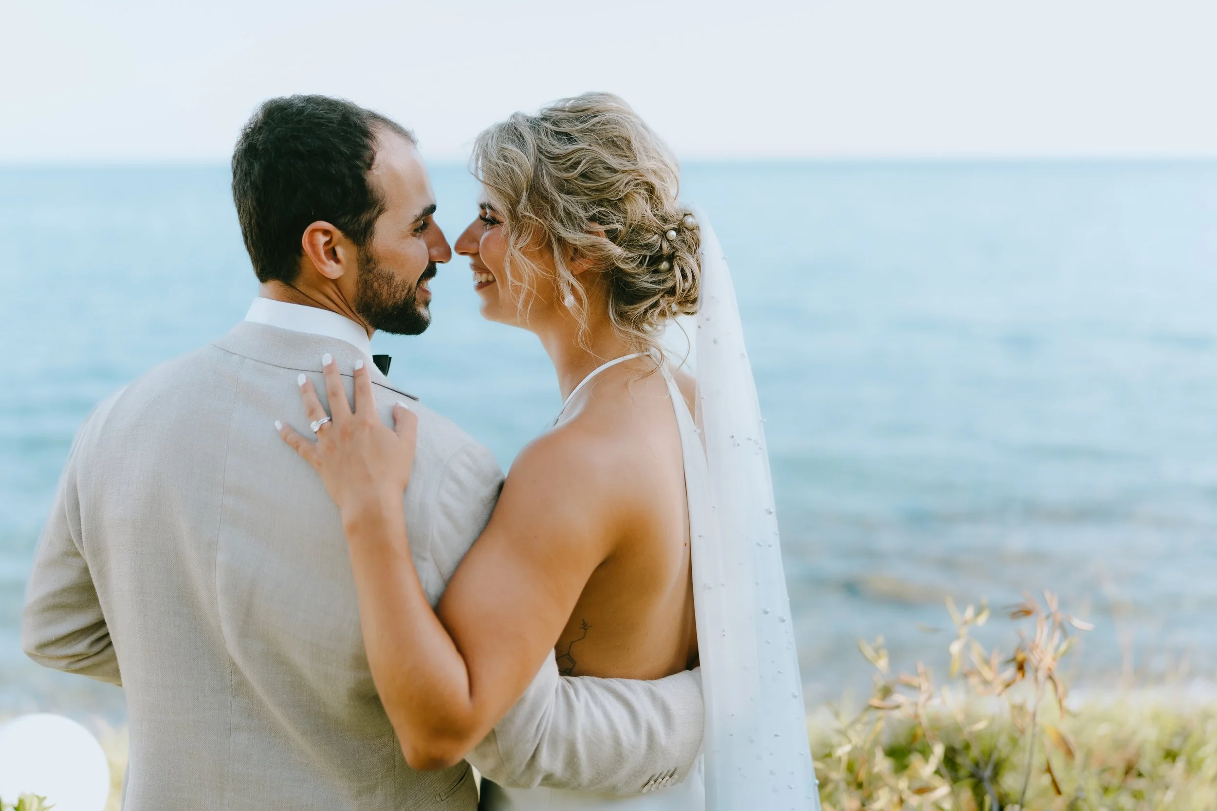 A bride and groom embrace on a beach, facing each other with their noses and foreheads touching, smiling at each other.