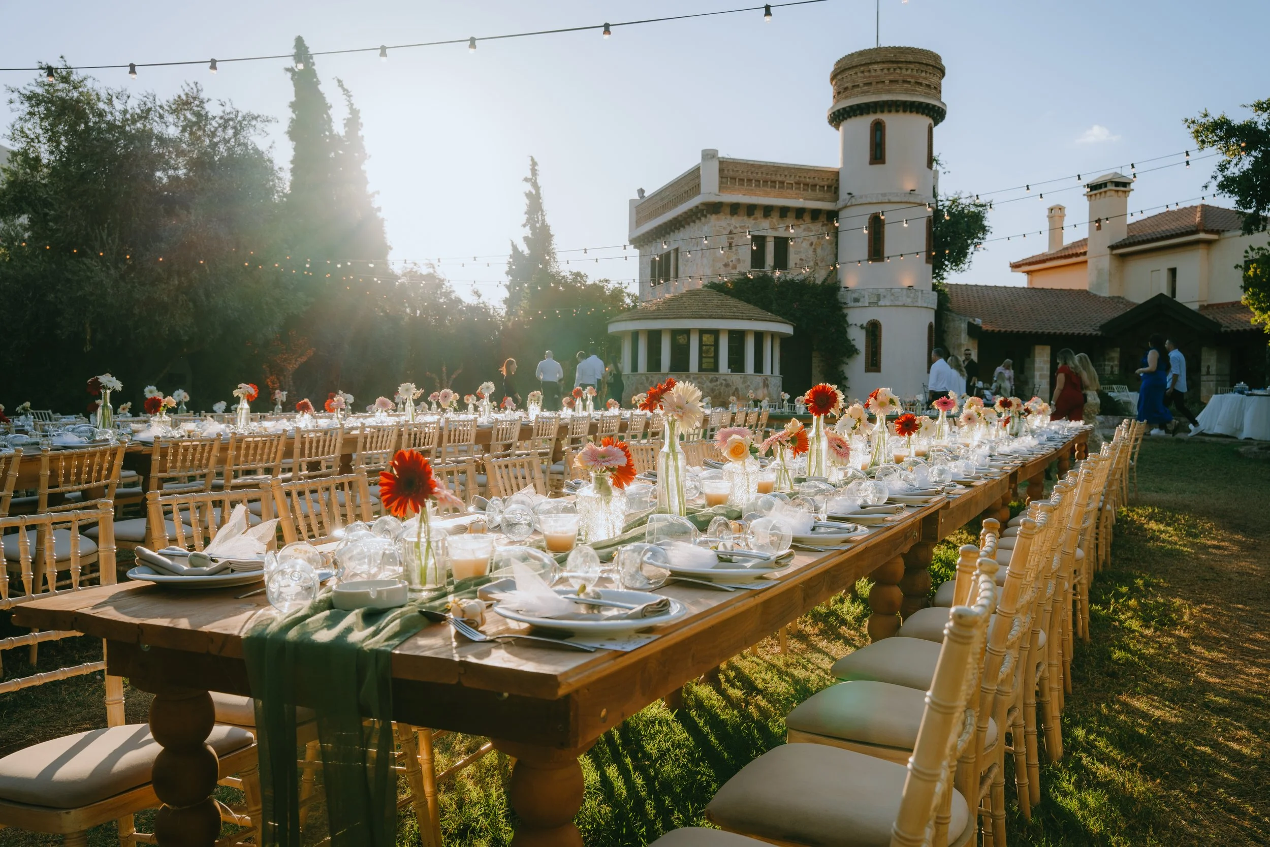 Outdoor banquet setup with long wooden table and chairs, decorated with vases of colorful flowers, in front of a castle-style building under string lights at sunset.