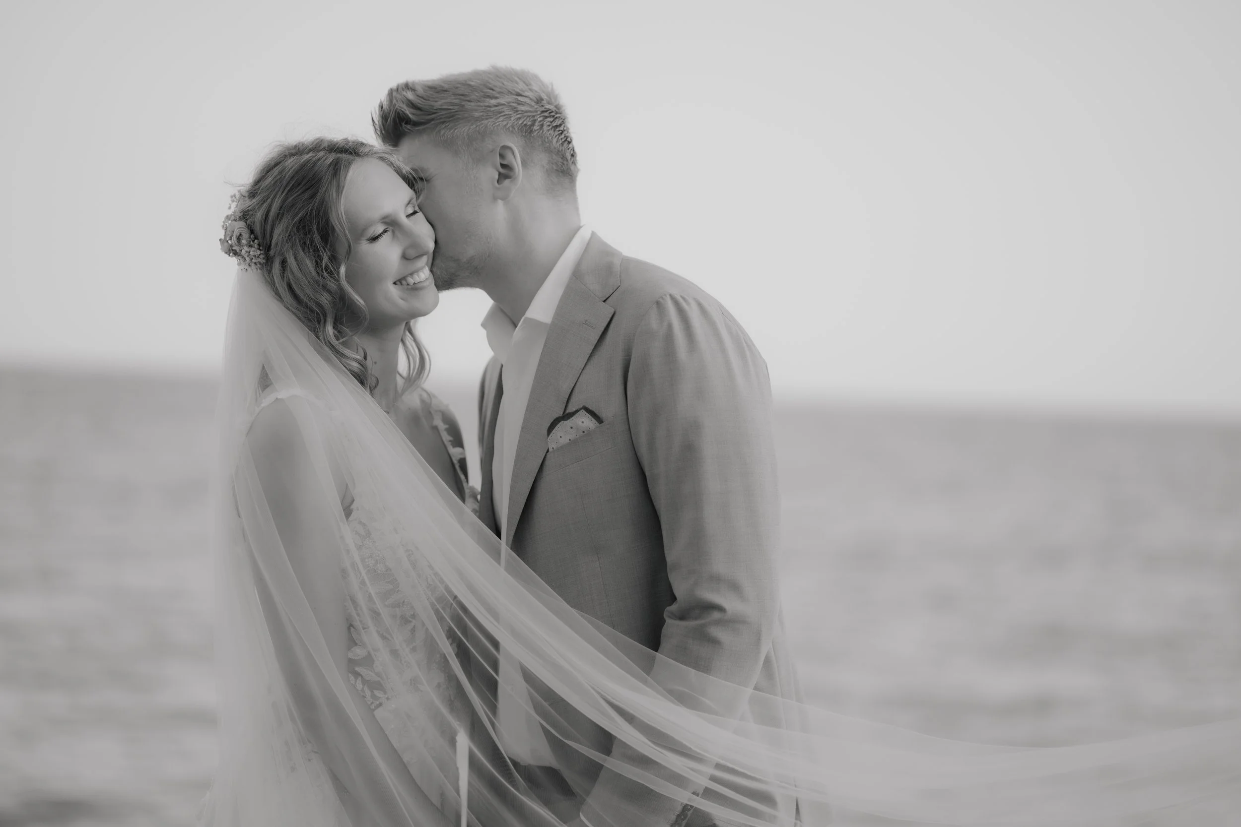 Black and white photo of a bride and groom smiling and embracing by the sea, the groom kissing the bride on her temple.