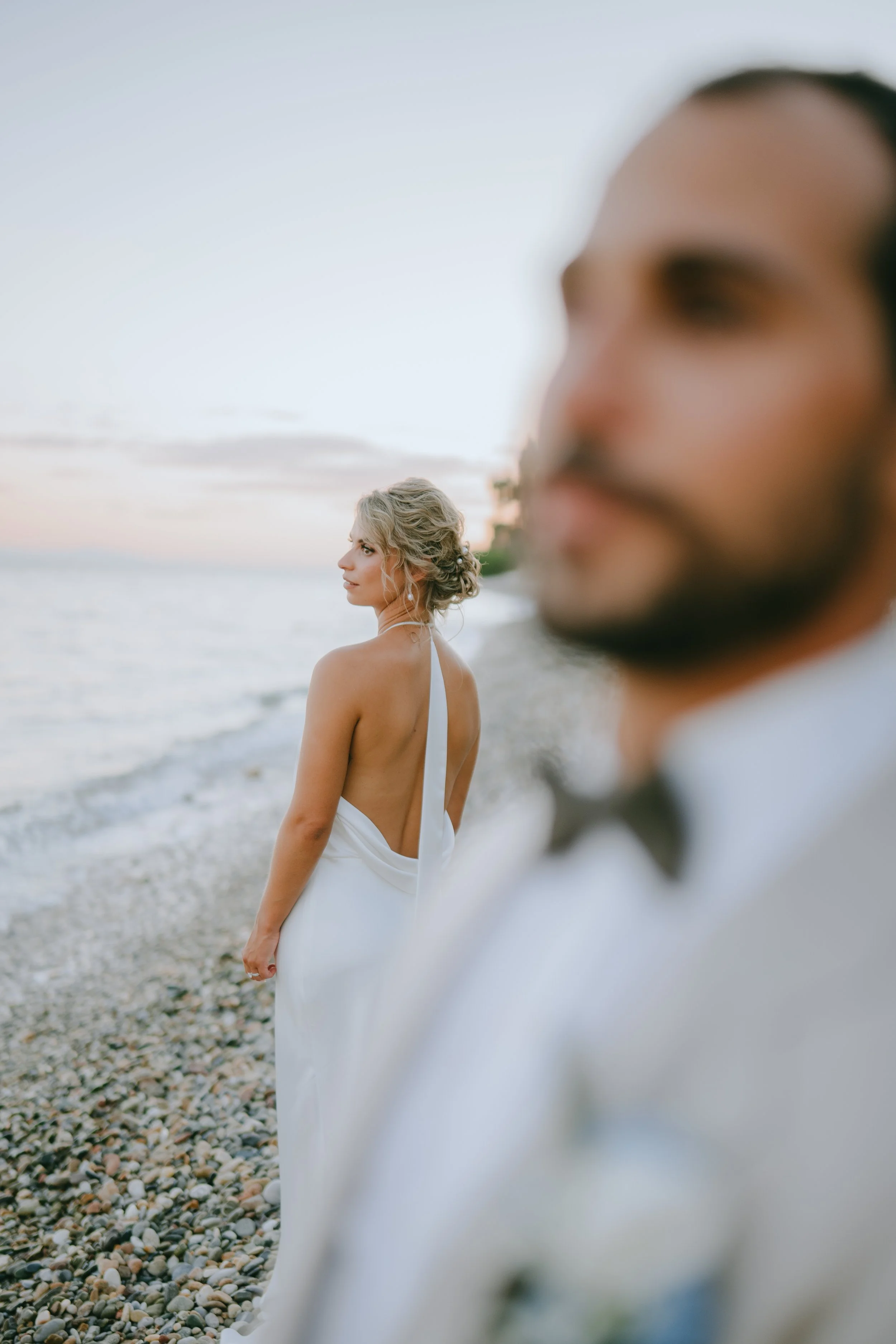A woman in a white dress standing on a pebbled beach near the ocean, with a man in a tuxedo blurred in the foreground.