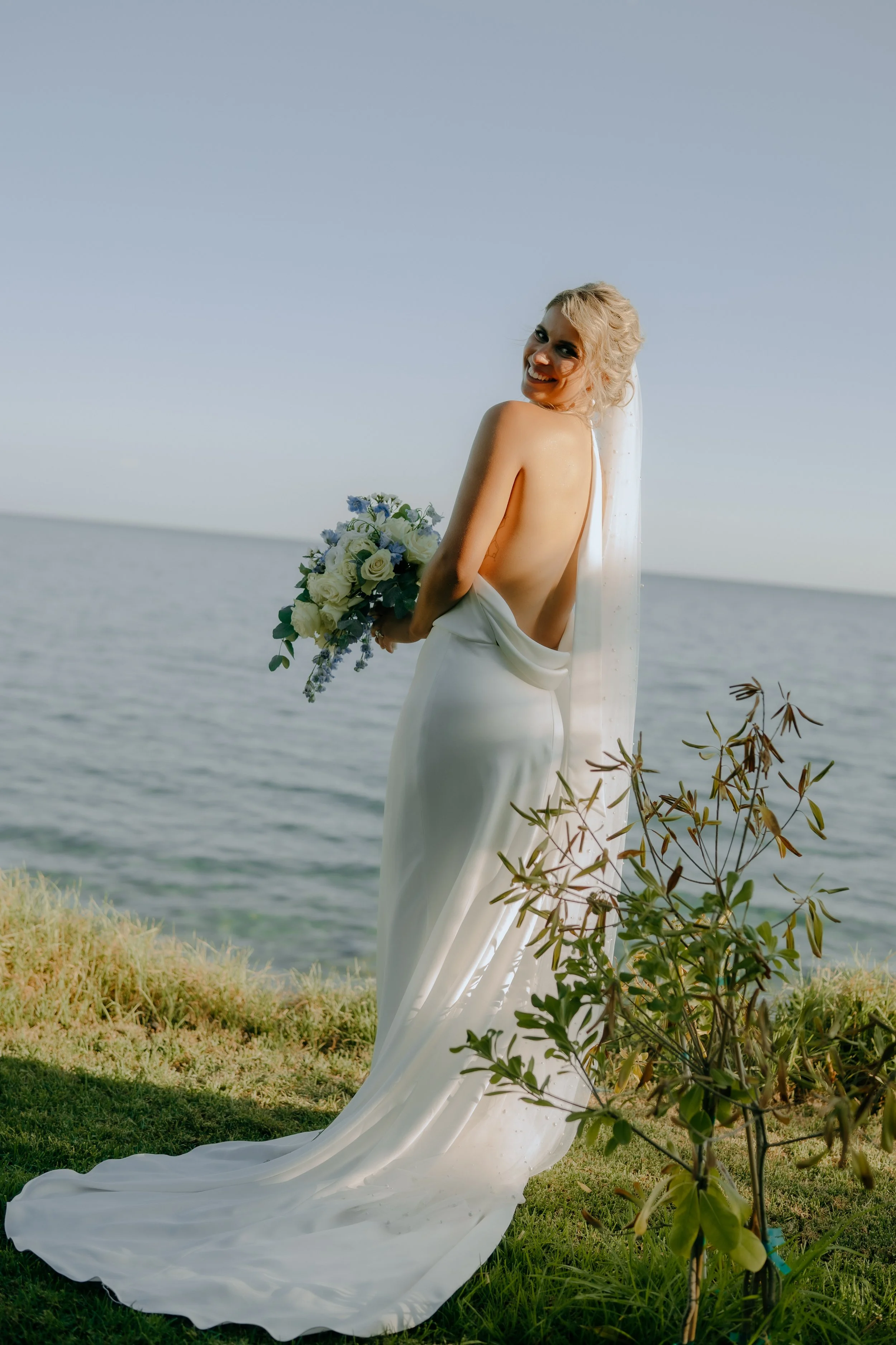 A woman in a white wedding dress with a long train holding a bouquet of white and purple flowers standing on a grassy area near the ocean, smiling at the camera with her back partially exposed.