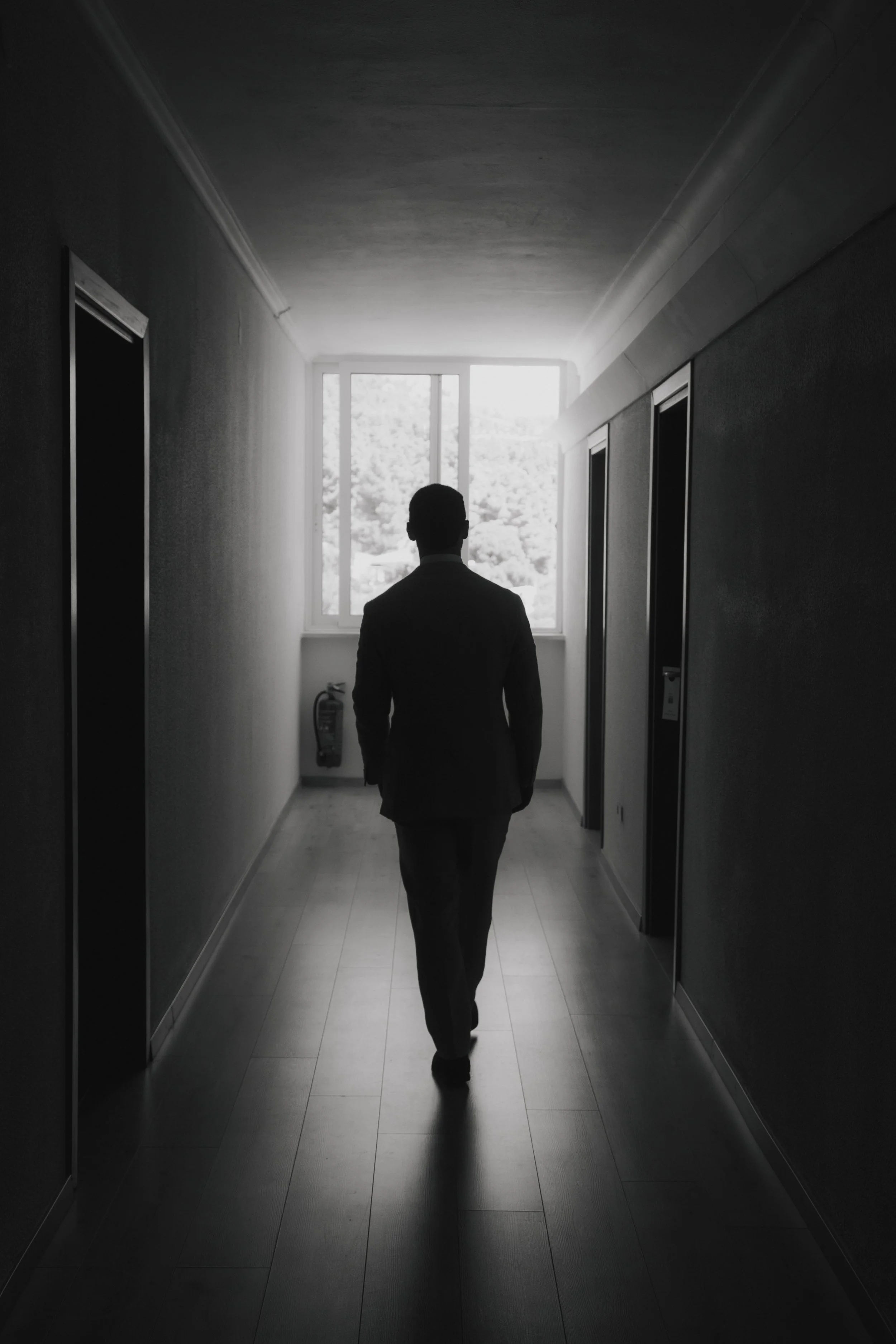A man in a suit walking down a dimly lit corridor towards a bright window.