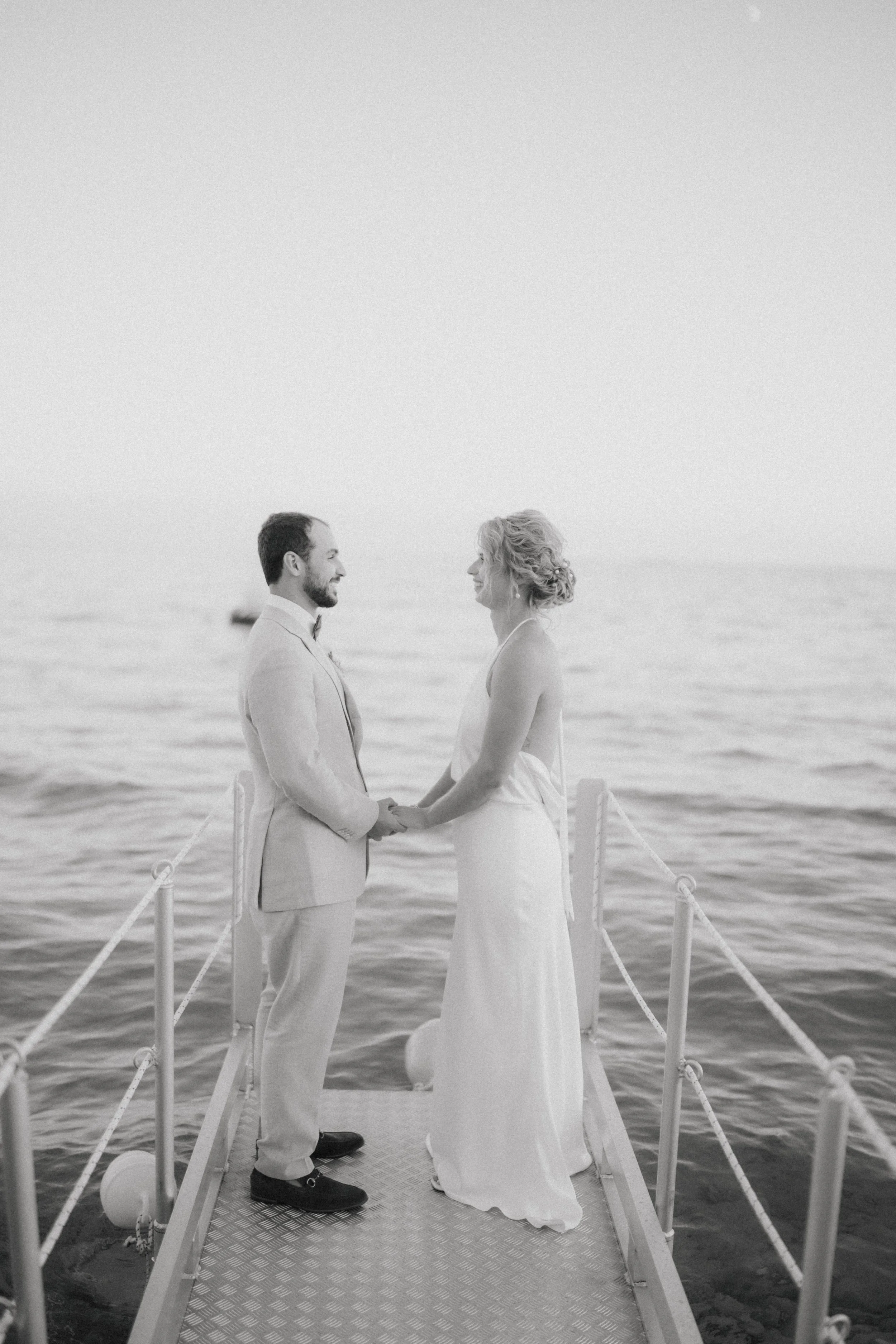 A black and white photograph of a wedding ceremony taking place on a boat docked in the water, featuring a bride and groom holding hands and smiling at each other with the sea in the background.