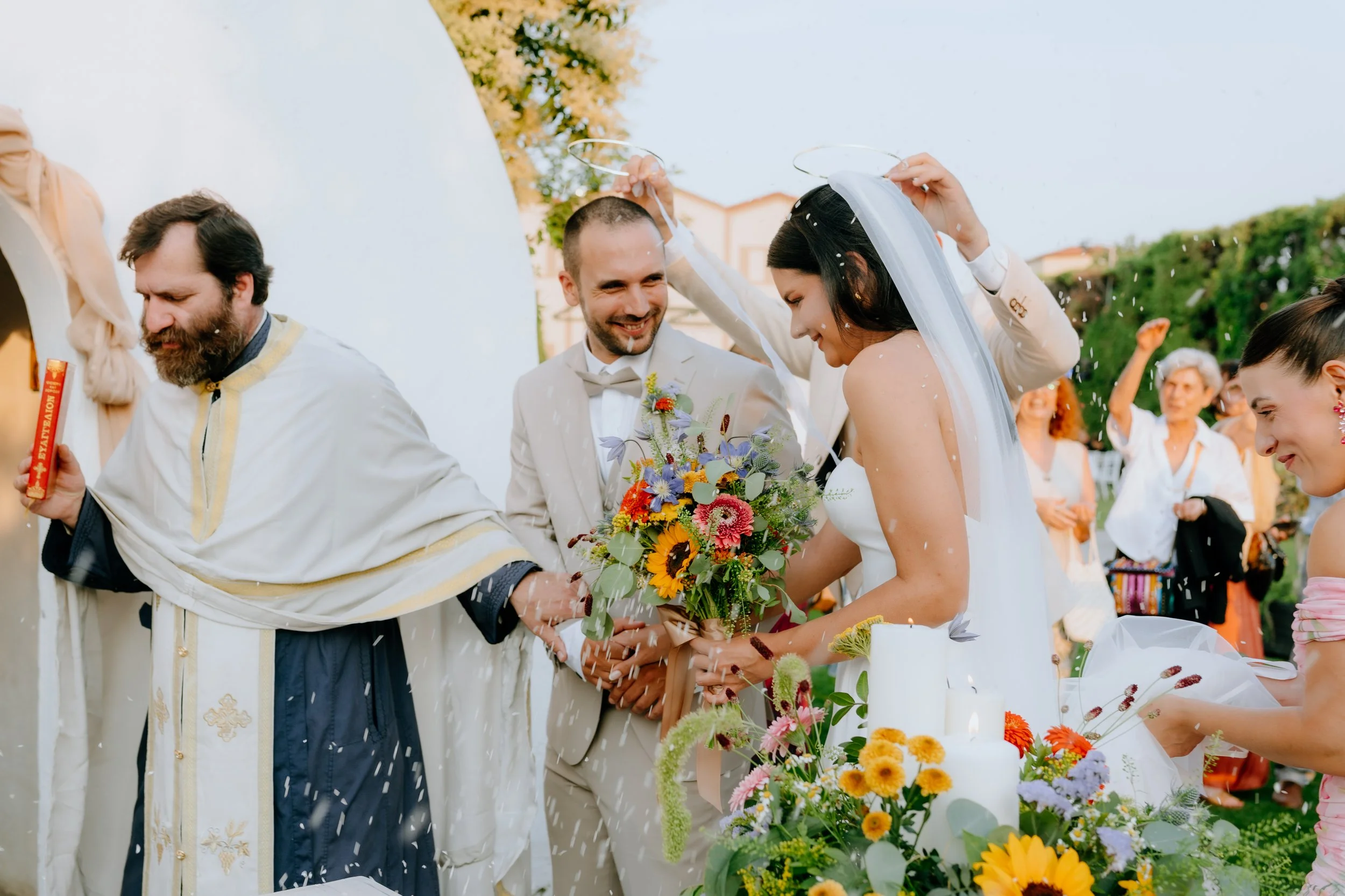 A wedding celebration outdoors with a bride and groom holding a bouquet, surrounded by guests. The bride is wearing a white dress and veil, and the groom in a light-colored suit. People are smiling; some are throwing confetti or rice.
