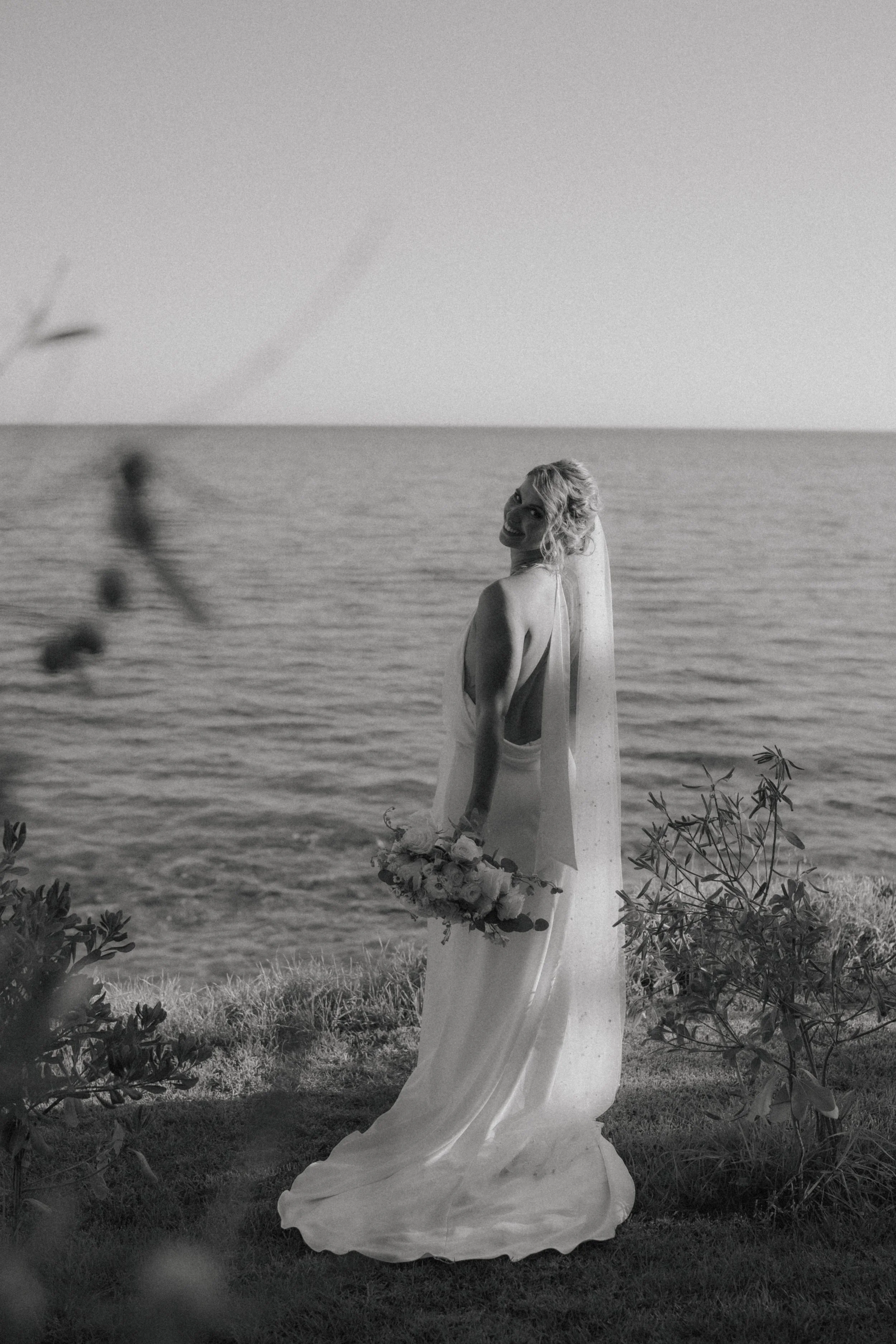 A bride in a sleeveless wedding dress holding a bouquet of flowers, standing outdoors near the ocean at sunset.