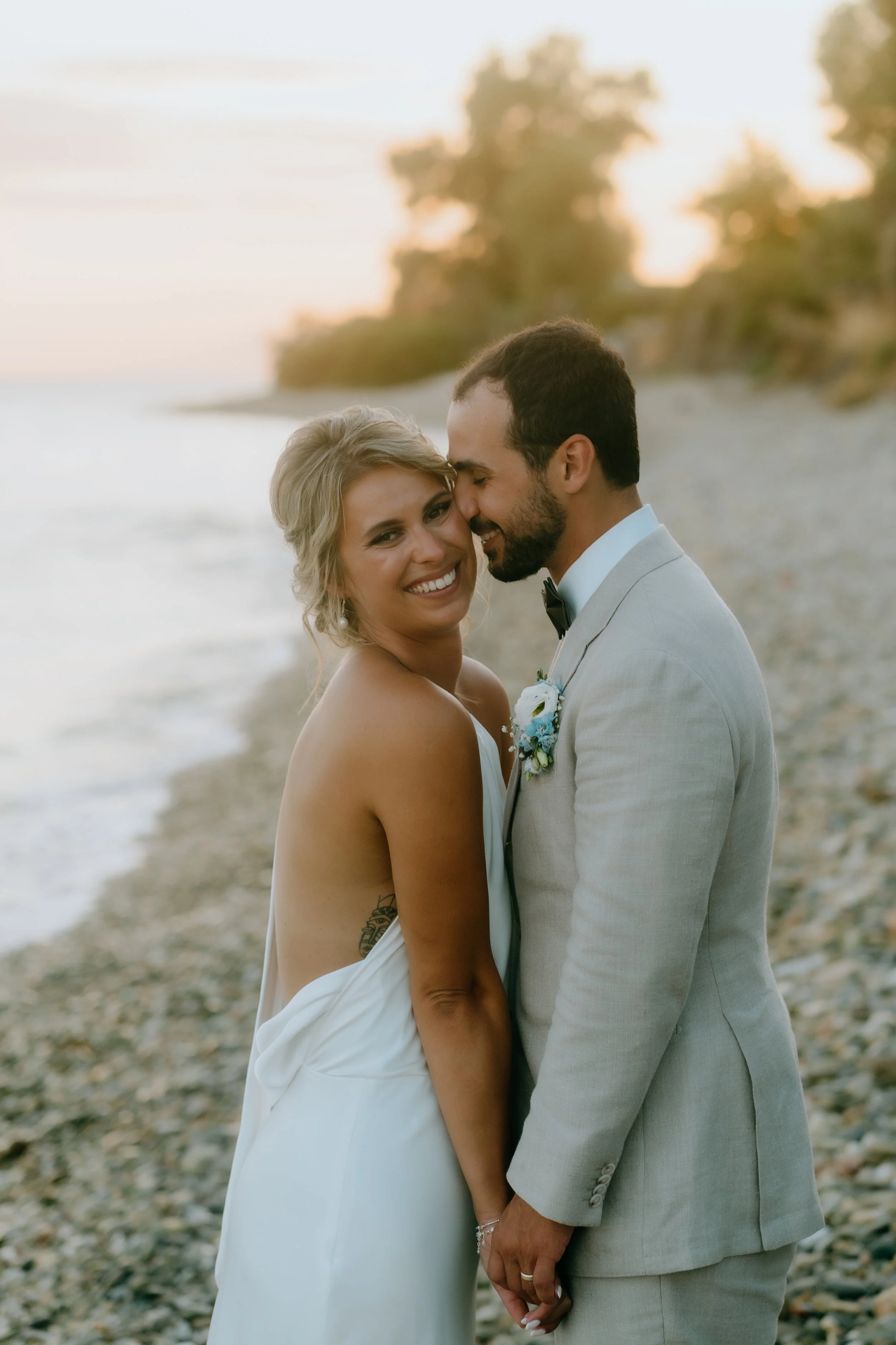 A newly married couple standing on a beach at sunset, smiling romantically. The woman is wearing a white dress with an open back, and the man is dressed in a light gray suit with a boutonnière. They are holding hands, with the woman smiling directly 