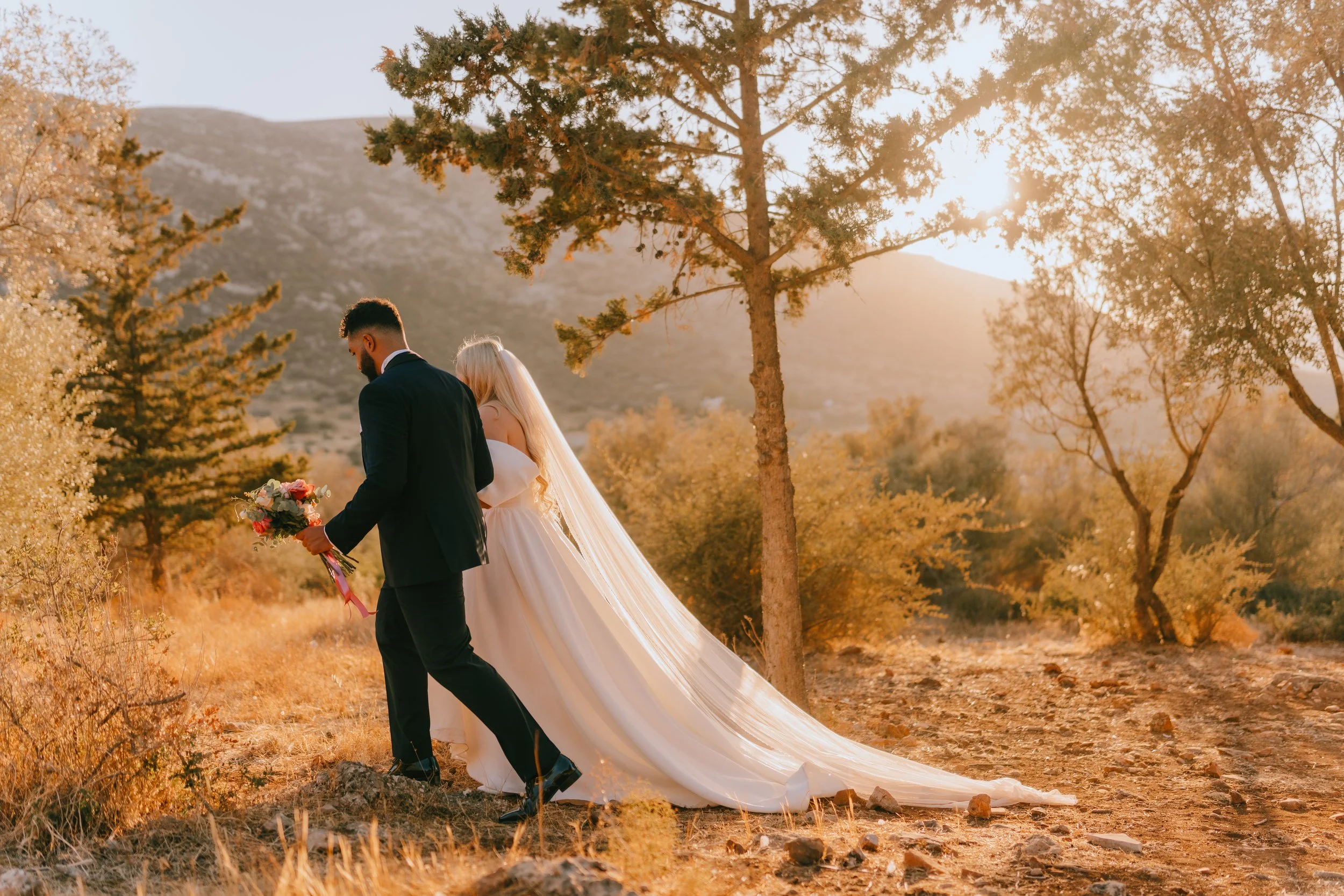 A bride and groom walking outdoors in a scenic, wooded area during sunset, with the bride wearing a long white wedding dress and veil, holding a bouquet of flowers, and the groom in a black suit.