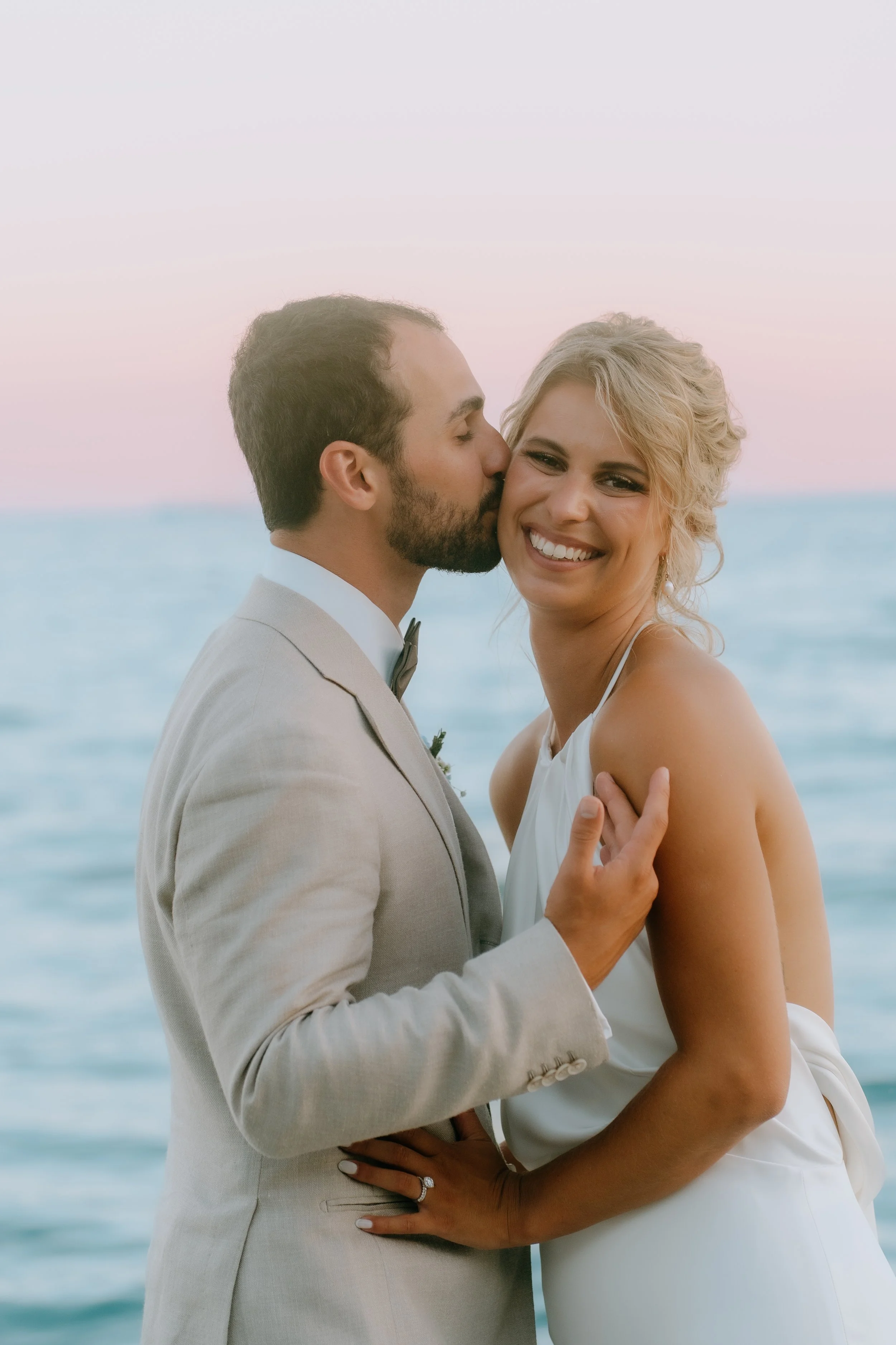 A newlywed couple stands close together outdoors with the ocean in the background; the man is kissing the woman's cheek, who is smiling.