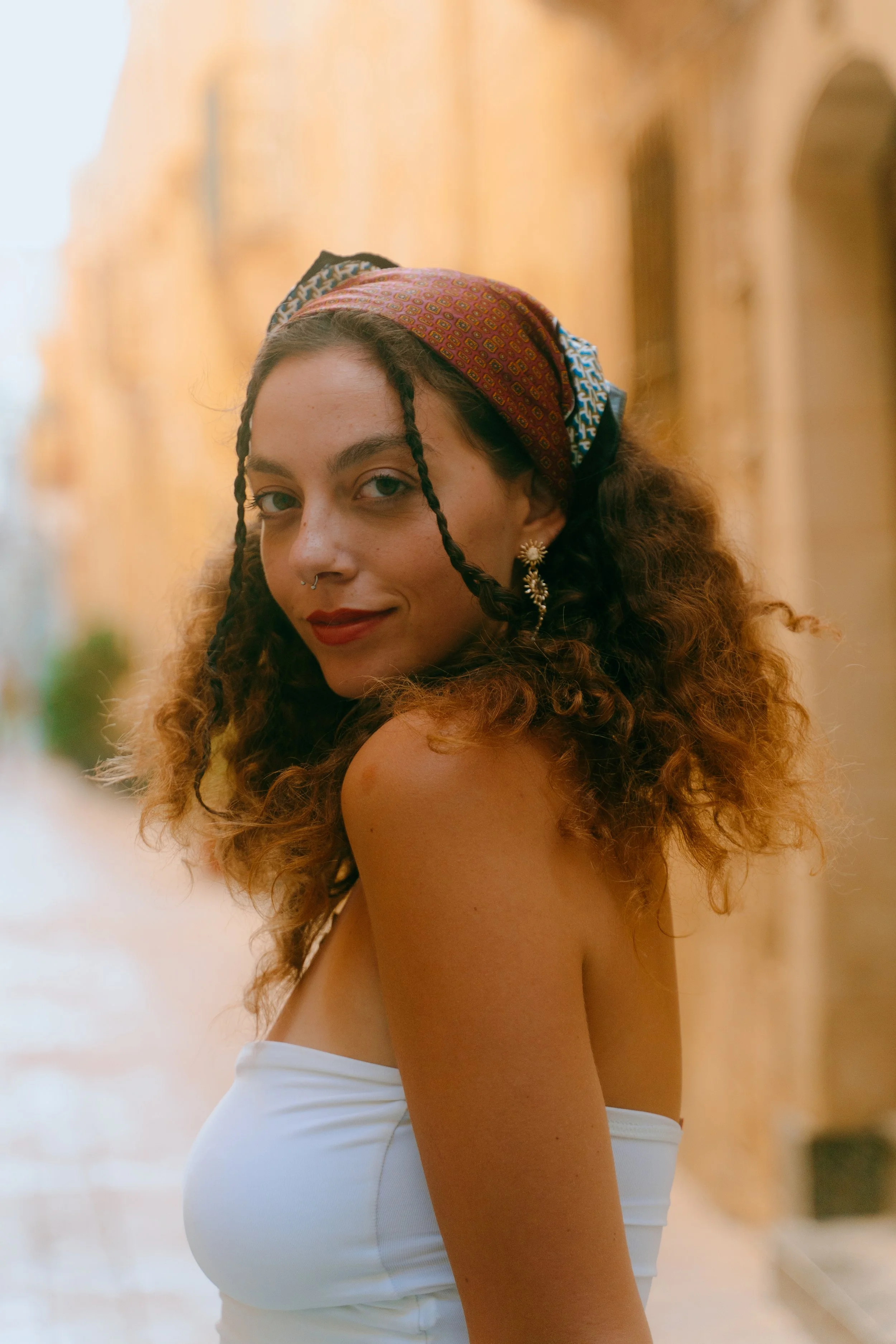 A young woman with curly hair and a colorful headscarf, wearing earrings and a white strapless top, standing outdoors with warm-colored buildings in the background.