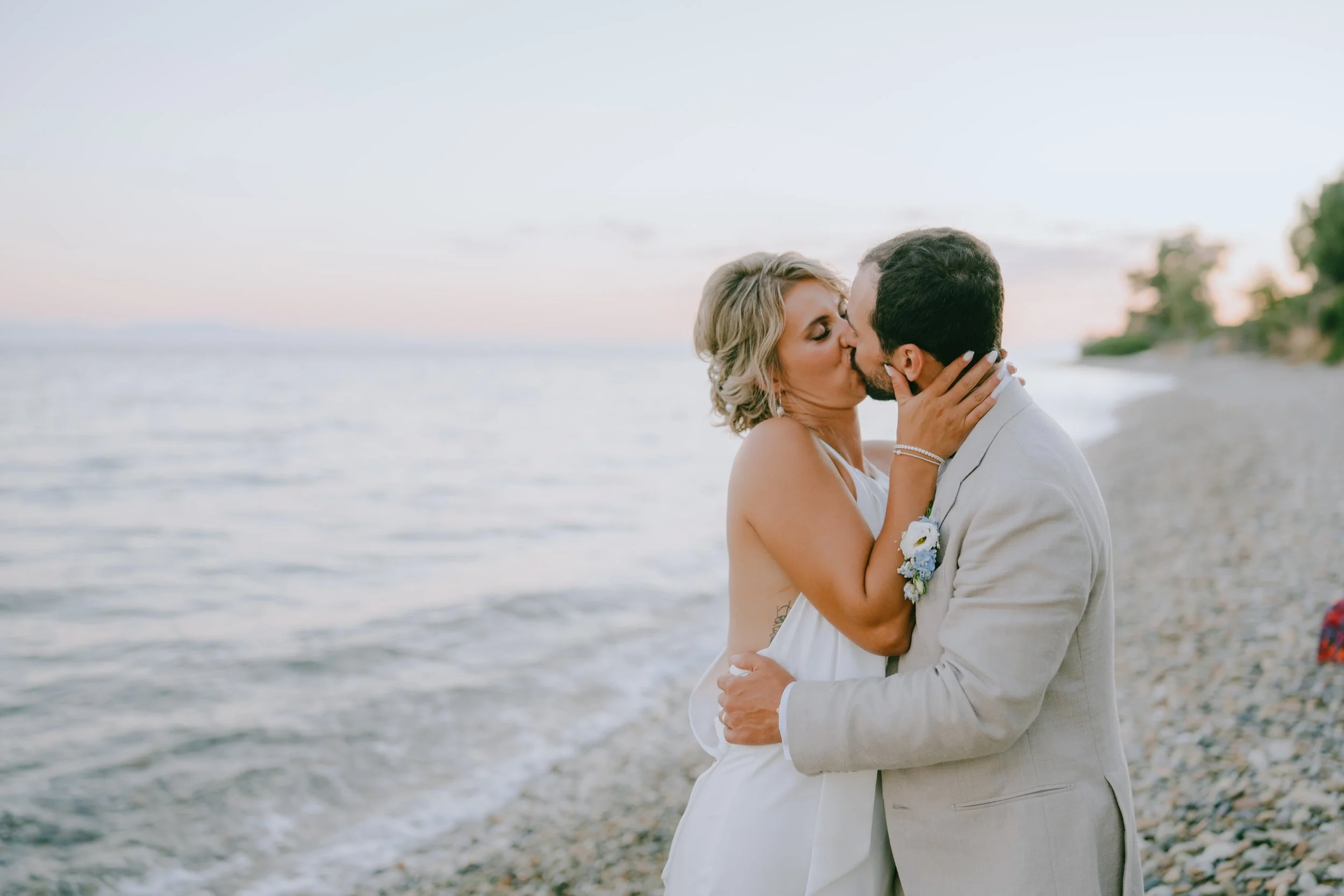 A couple kissing on a pebble beach at sunset, with the ocean and trees in the background.