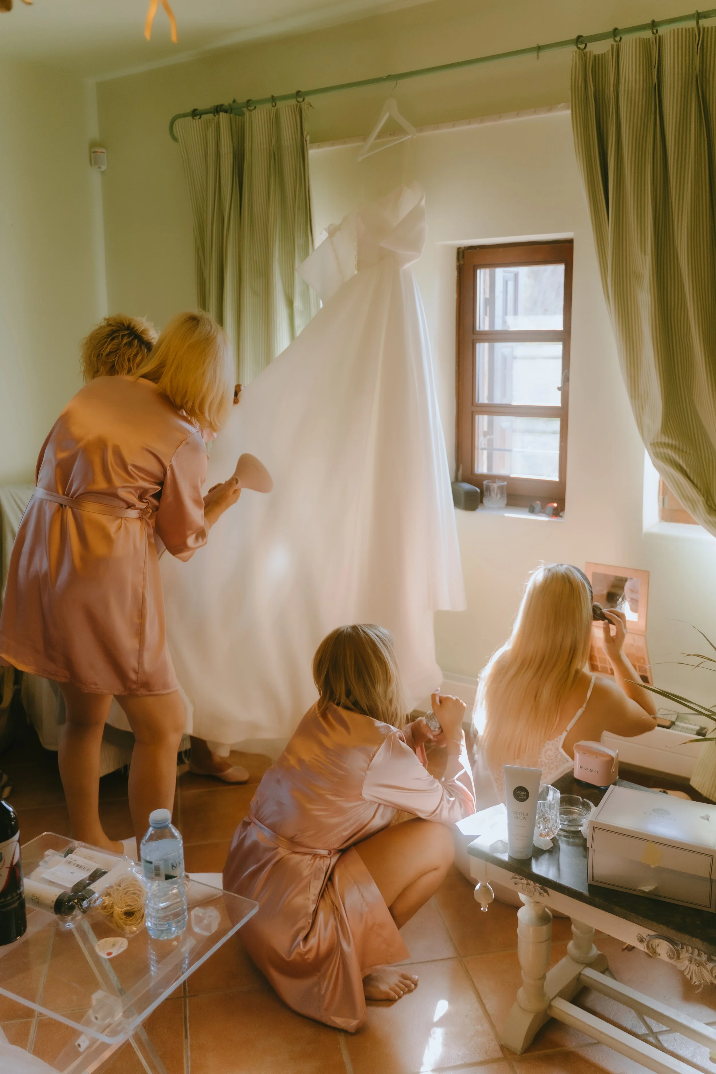 Group of women in pink satin robes preparing a white wedding dress hanging by the window in a bright room.