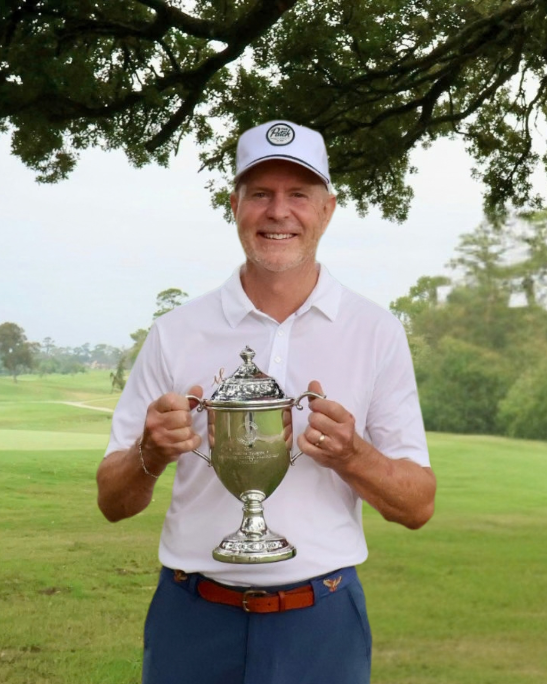 A man smiling and holding a large silver trophy on a golf course, wearing a white polo shirt, a white cap, and blue pants.
