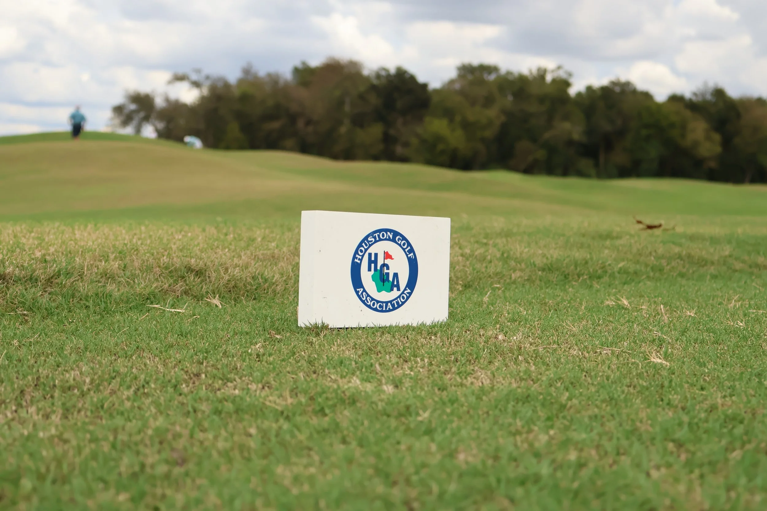 A golf course with a sign in the foreground that says 'Houston Golf Association.' A golfer is visible in the distance on the green, with trees and a partly cloudy sky in the background.