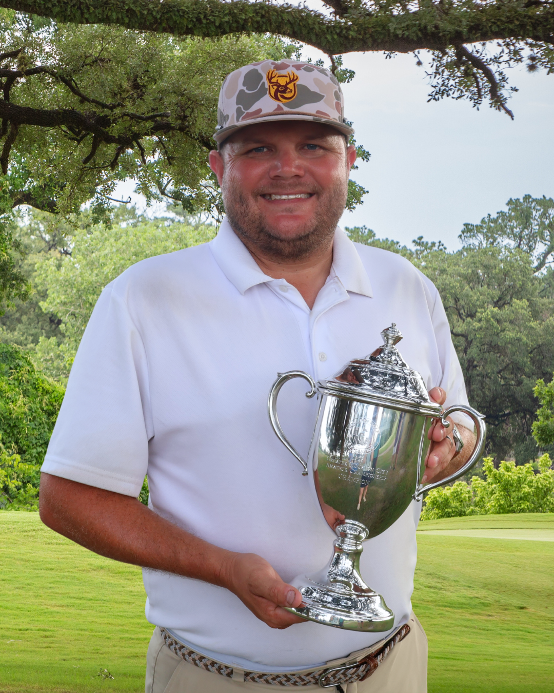 A man smiling and holding a silver trophy on a golf course surrounded by greenery and trees.