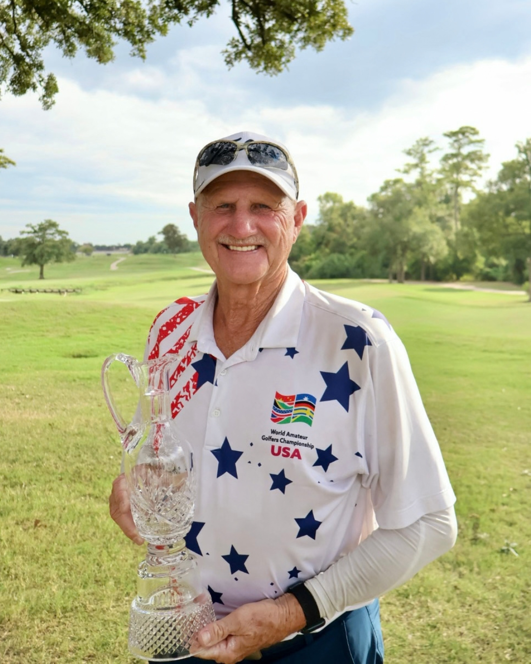 An older man holding a crystal trophy, dressed in a patriotic golf outfit, smiling on a golf course with trees and open sky in the background.