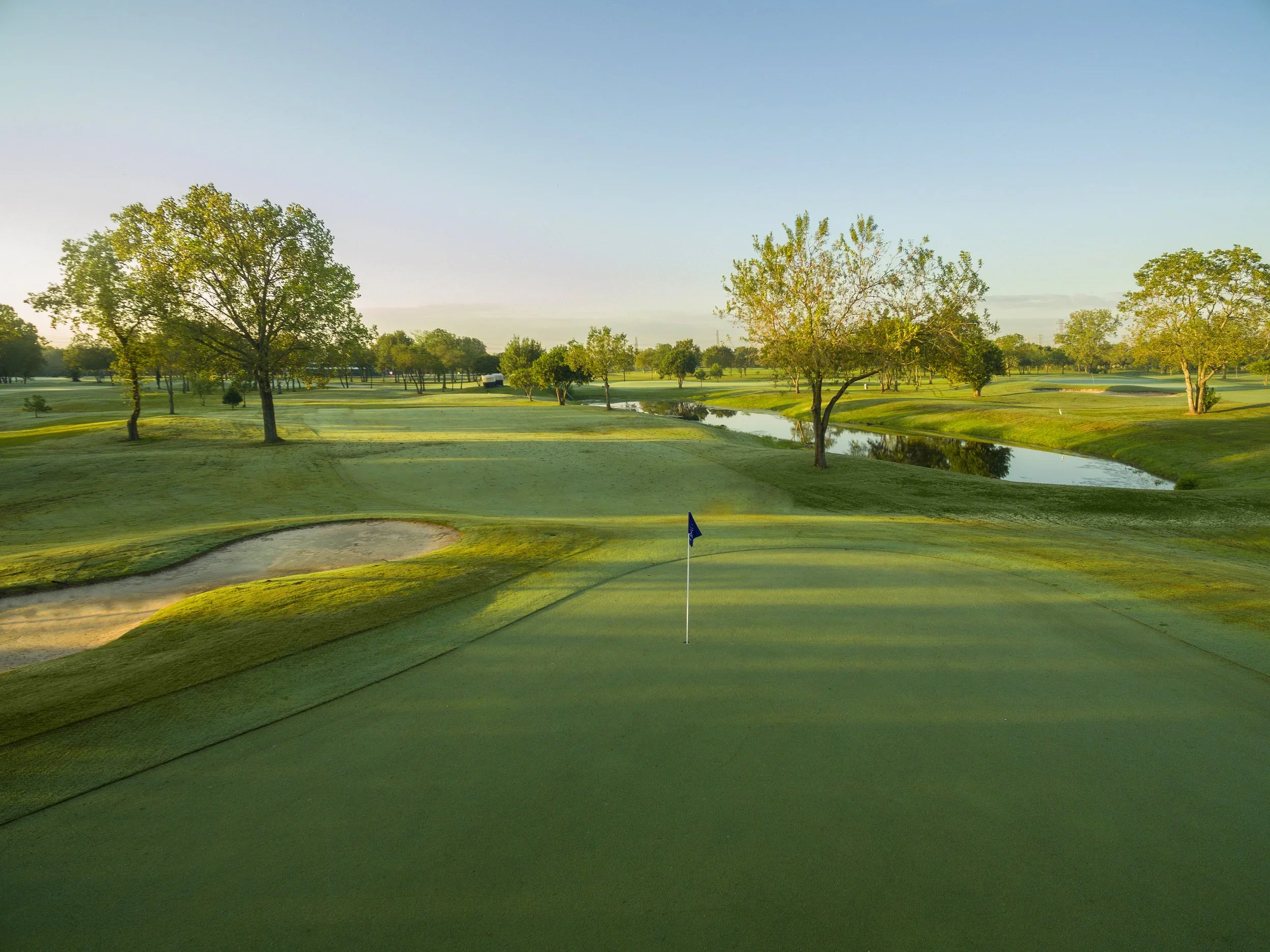 A golf course with neatly trimmed grass, a sand trap in the foreground, a small pond, and several trees under a clear blue sky during the daytime.