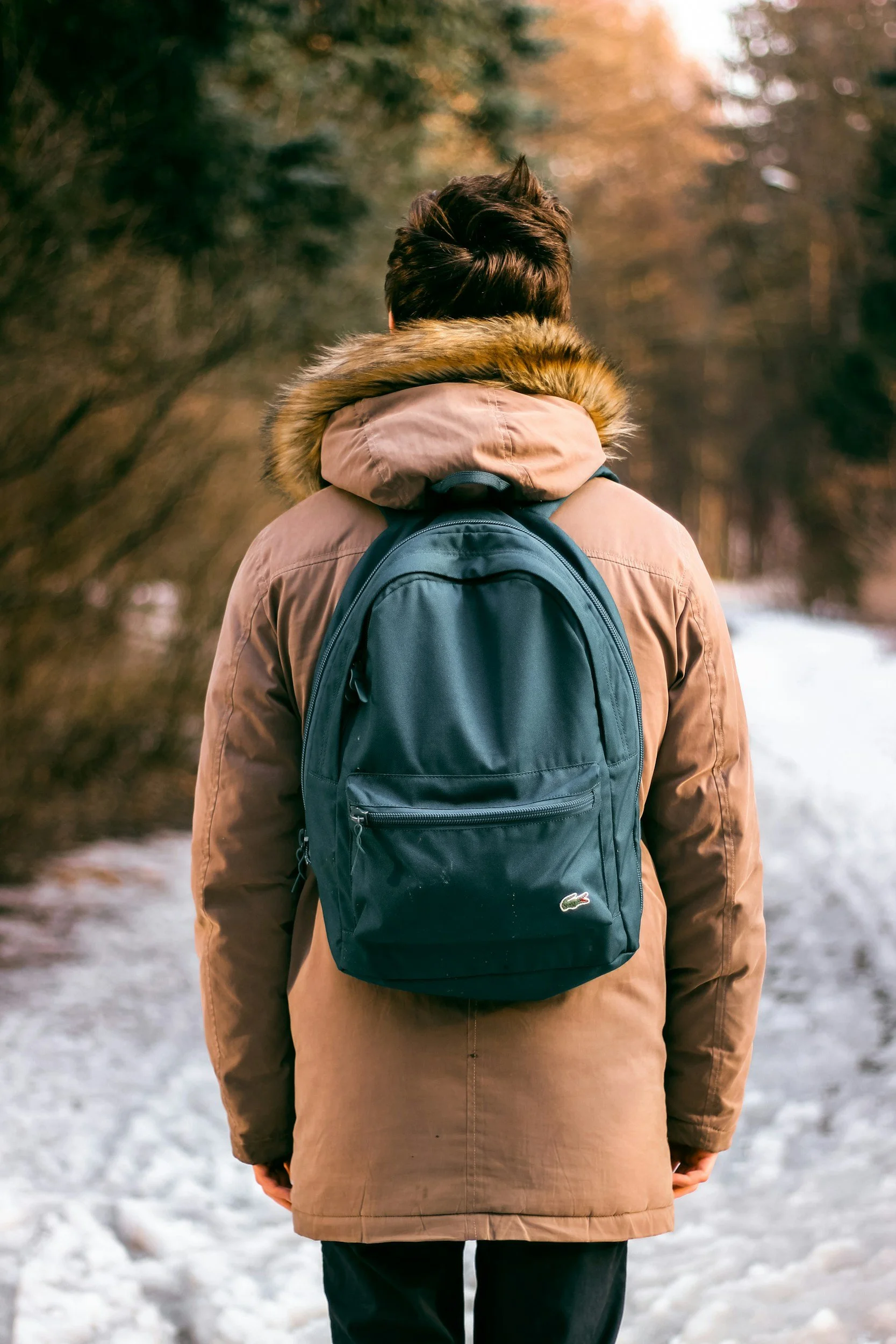 Person wearing a tan winter coat with a fur-lined hood and a dark blue backpack, walking on a snow-covered path in a forest during autumn.