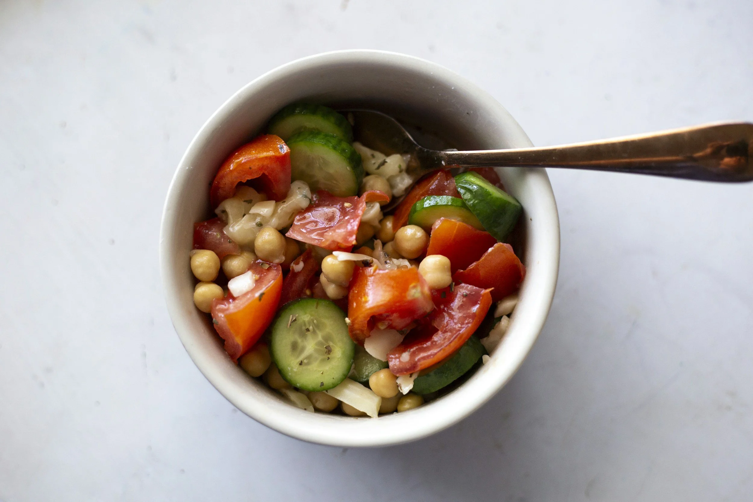 A white bowl filled with a vegetable salad containing sliced cucumbers, cherry tomatoes, chickpeas, and shredded cheese, with a spoon resting inside.