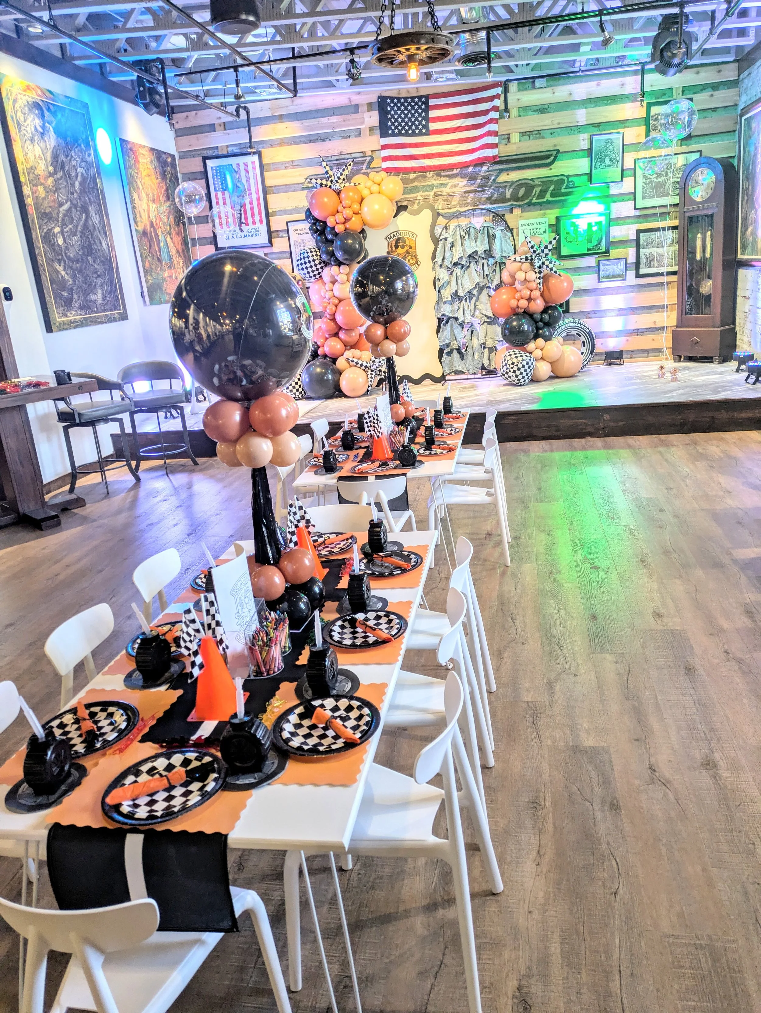 Decorated event space with a racing theme, featuring black, orange, and checkered balloons, checkered table settings, and a stage with a vintage grandfather clock and framed photos, along with an American flag on the wall.