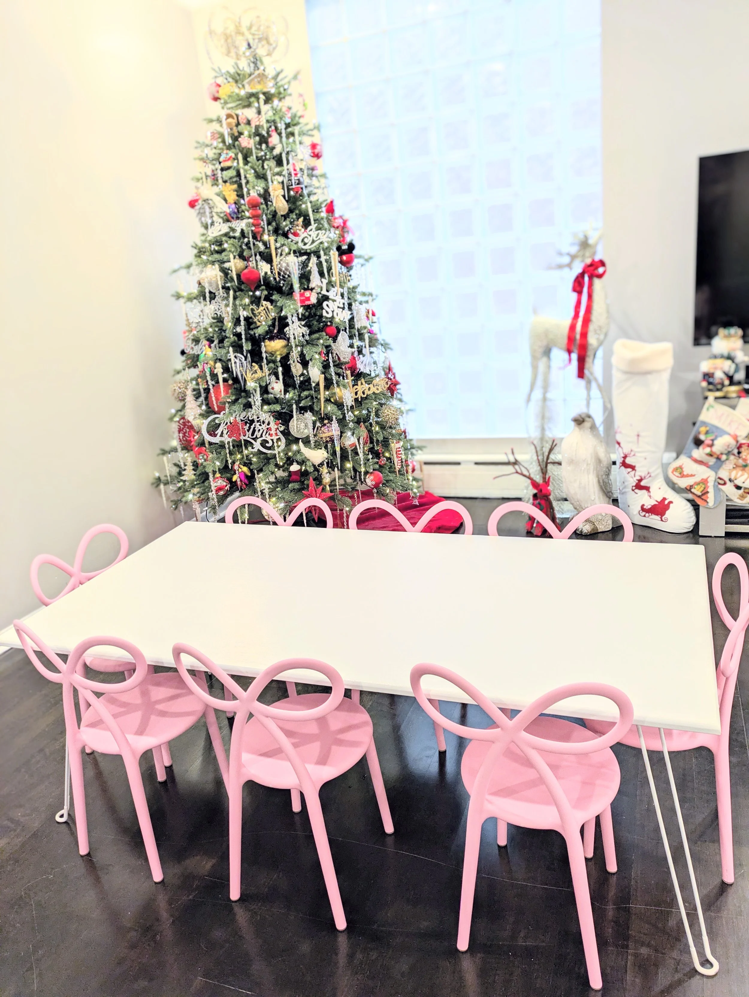 A room decorated for Christmas with a large decorated Christmas tree, a white reindeer ornament with a red bow, and a white table surrounded by pink chairs with heart-shaped backs.
