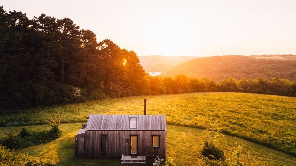 A small wooden cabin surrounded by green fields and trees at sunset.